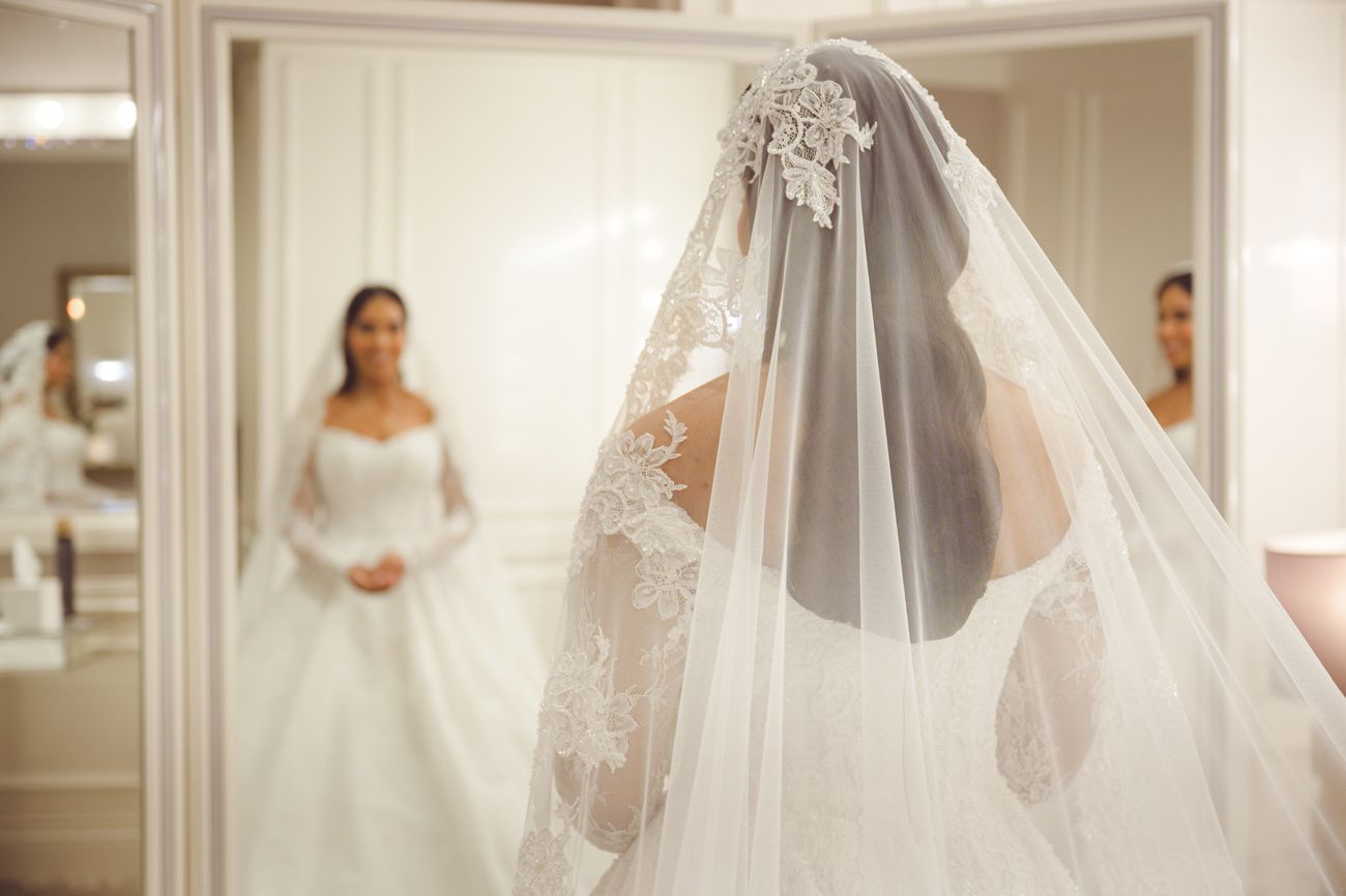 Bride in elegant white lace wedding gown with cathedral veil reflected in mirror at bridal boutique.