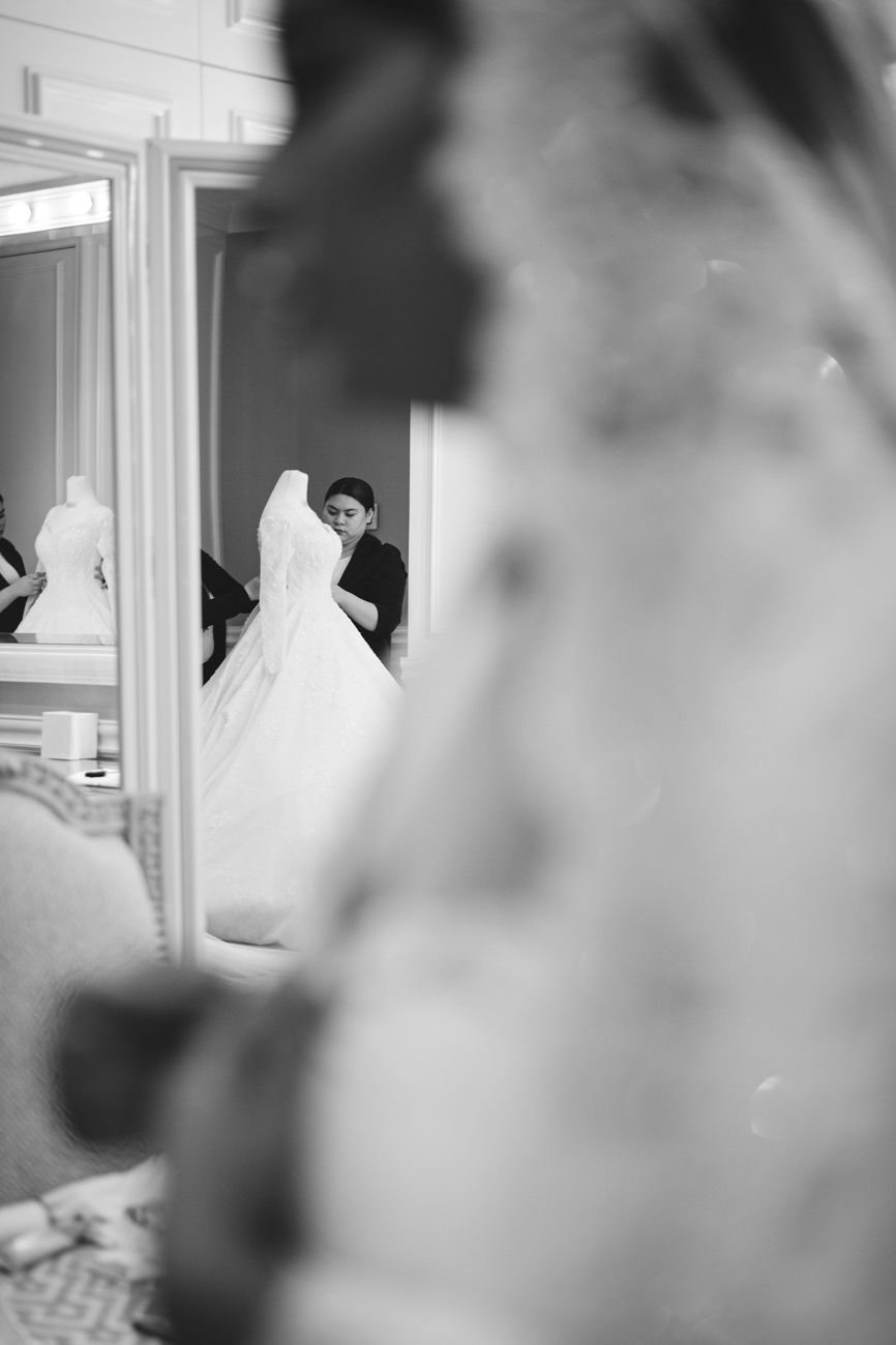 Black and white photo of a bride in a white gown being assisted by a dresser, reflected in a mirror.
