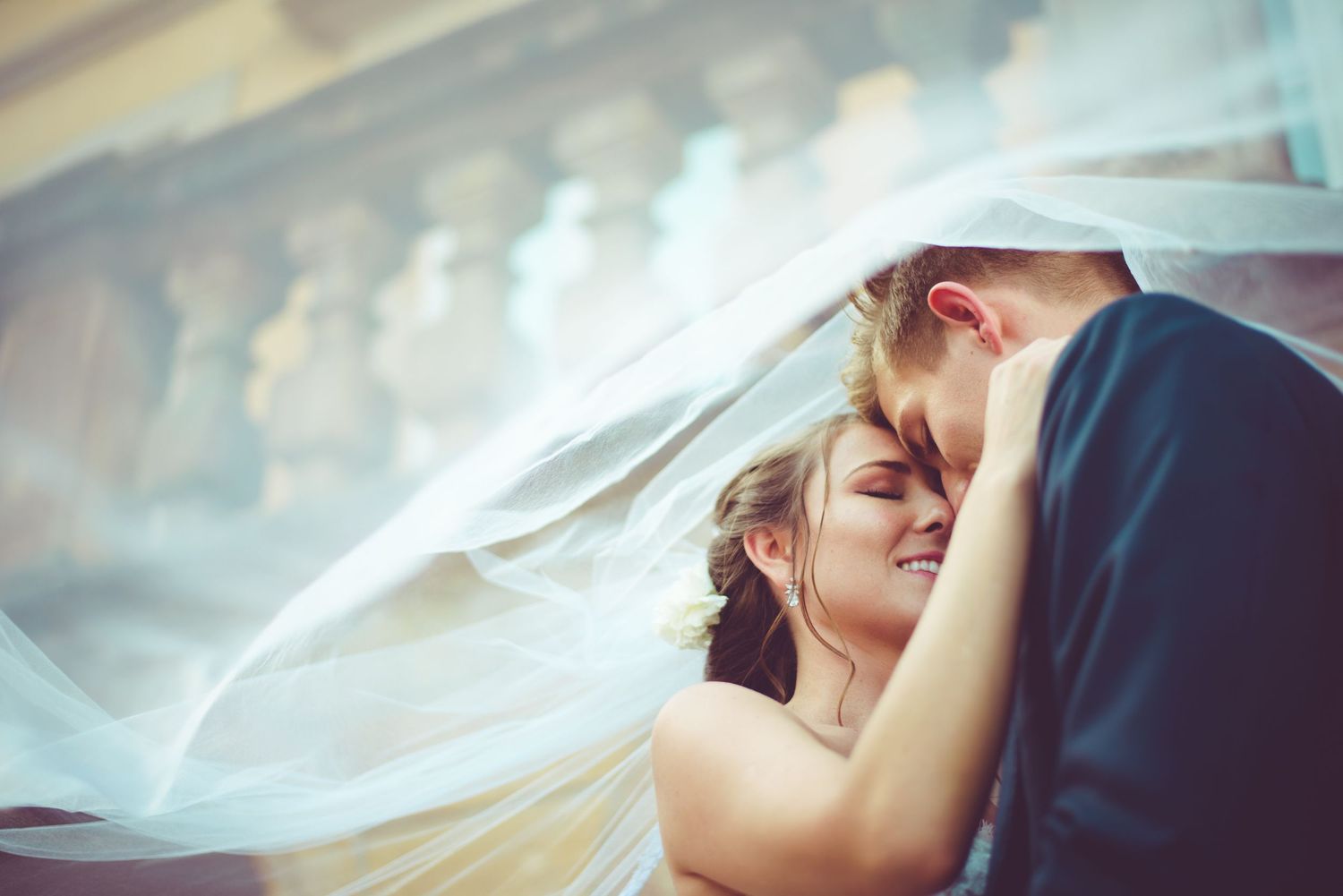 A bride and groom share an intimate moment under a flowing white wedding veil outdoors.