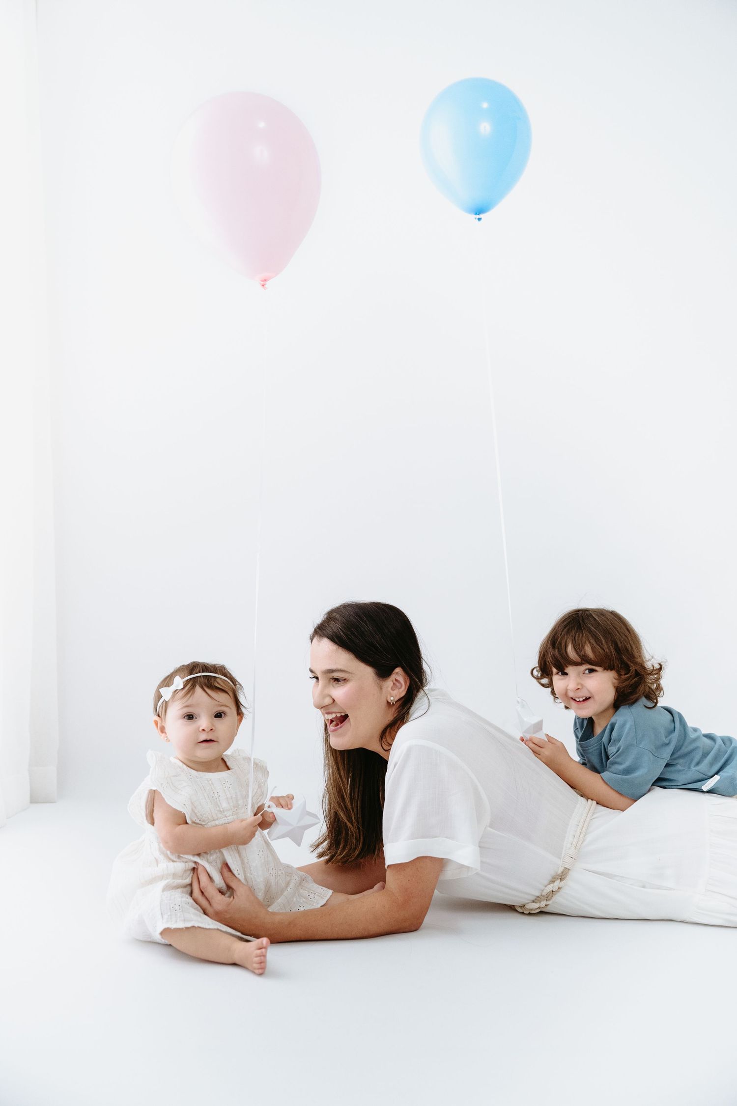 Mother lying on white floor with baby girl and toddler boy holding pink and blue balloons in bright studio.