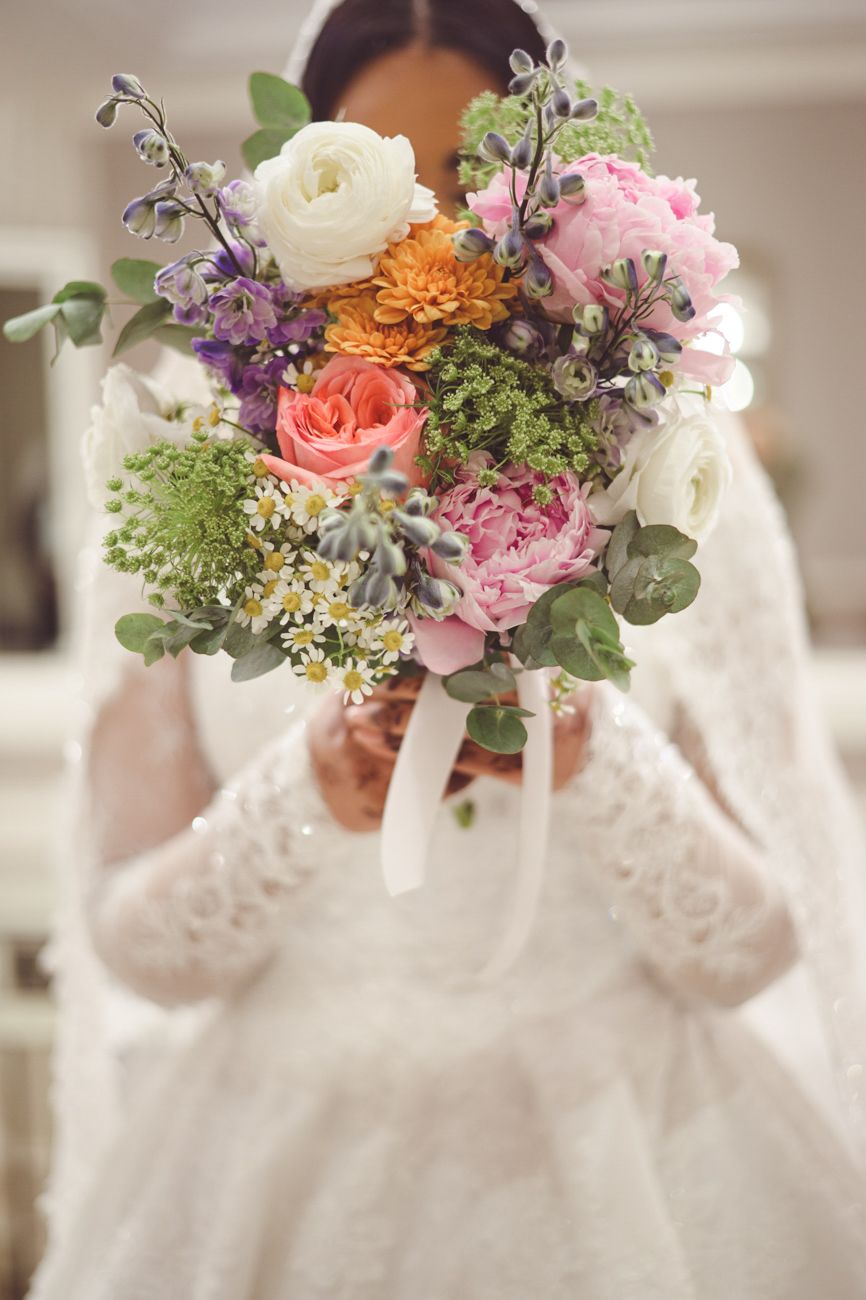 Bride in white lace gown holding colorful wedding bouquet with pink roses, orange dahlias, and purple blooms.
