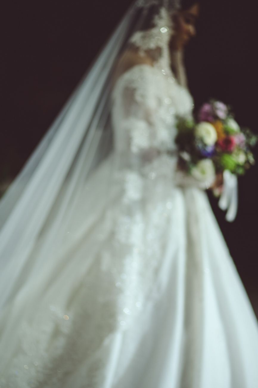 Blurred bride in white lace ballgown wedding dress with veil holding colorful bouquet against dark background.