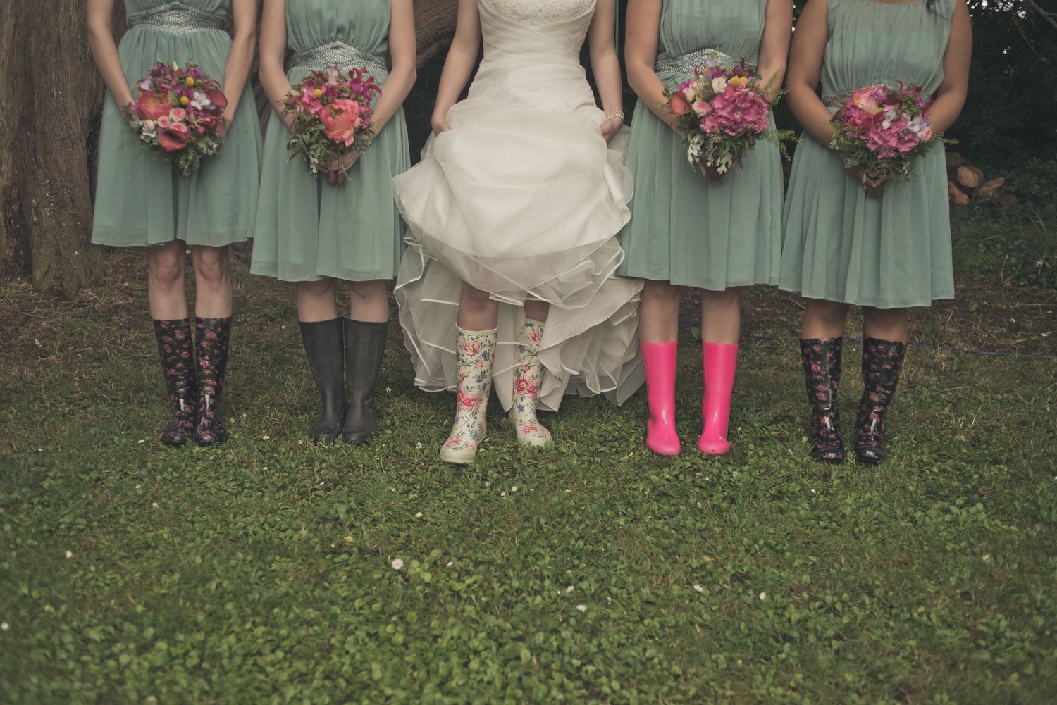 Bride and bridesmaids in mint dresses wearing colorful rain boots holding floral bouquets on green grass.