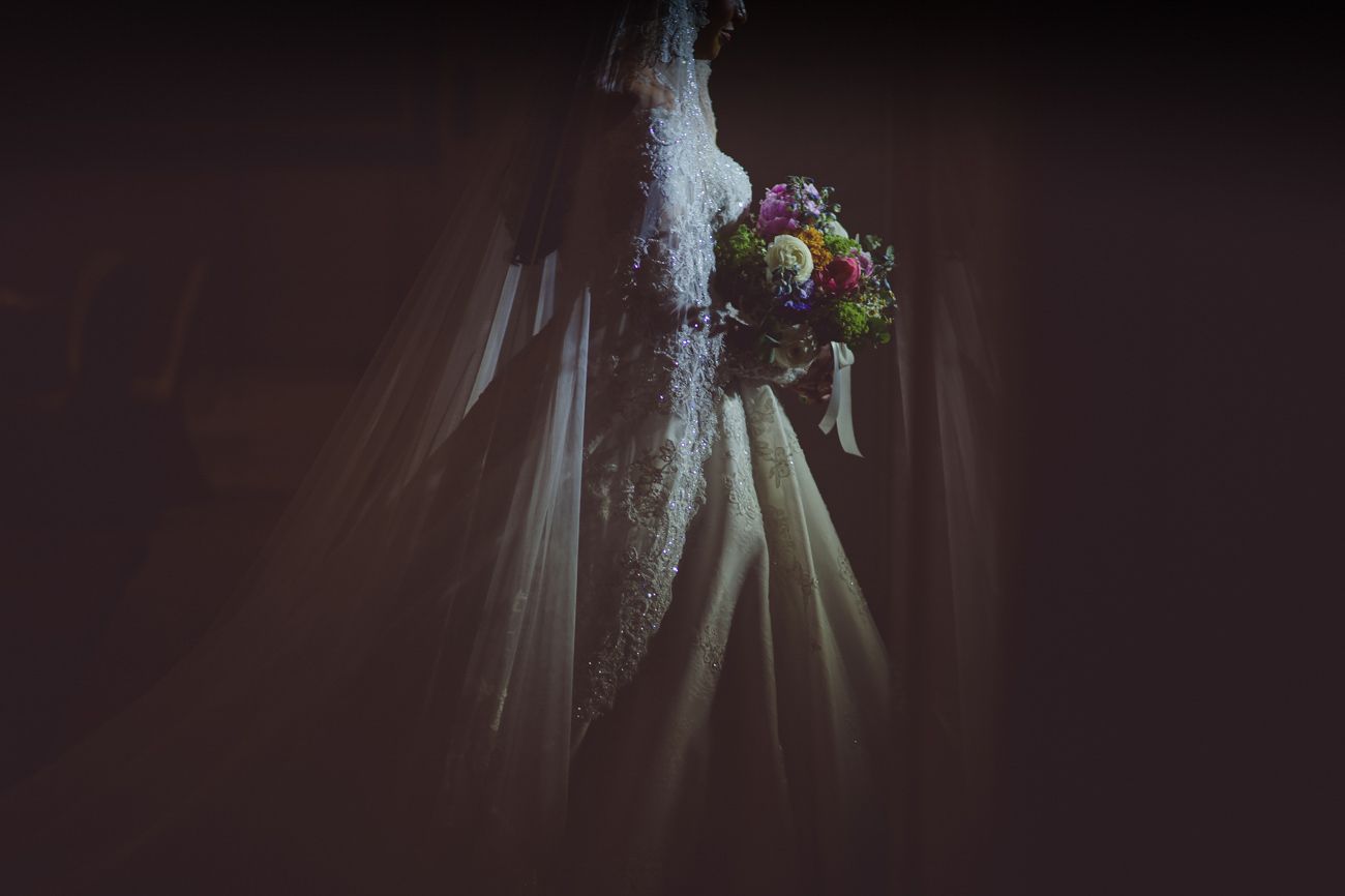 Bride in elegant lace wedding gown holding colorful bouquet with flowing veil in dramatic dark setting.