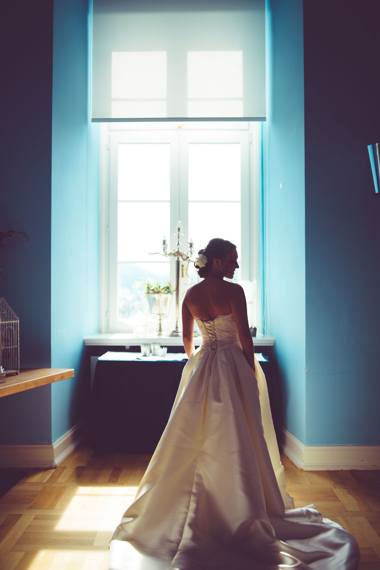 Bride in elegant white gown standing by sunlit window in blue room, silhouetted against bright natural light.