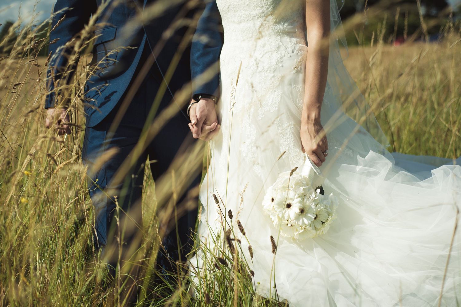 Bride in white lace gown holding bouquet stands with groom in field of tall grass during outdoor wedding.