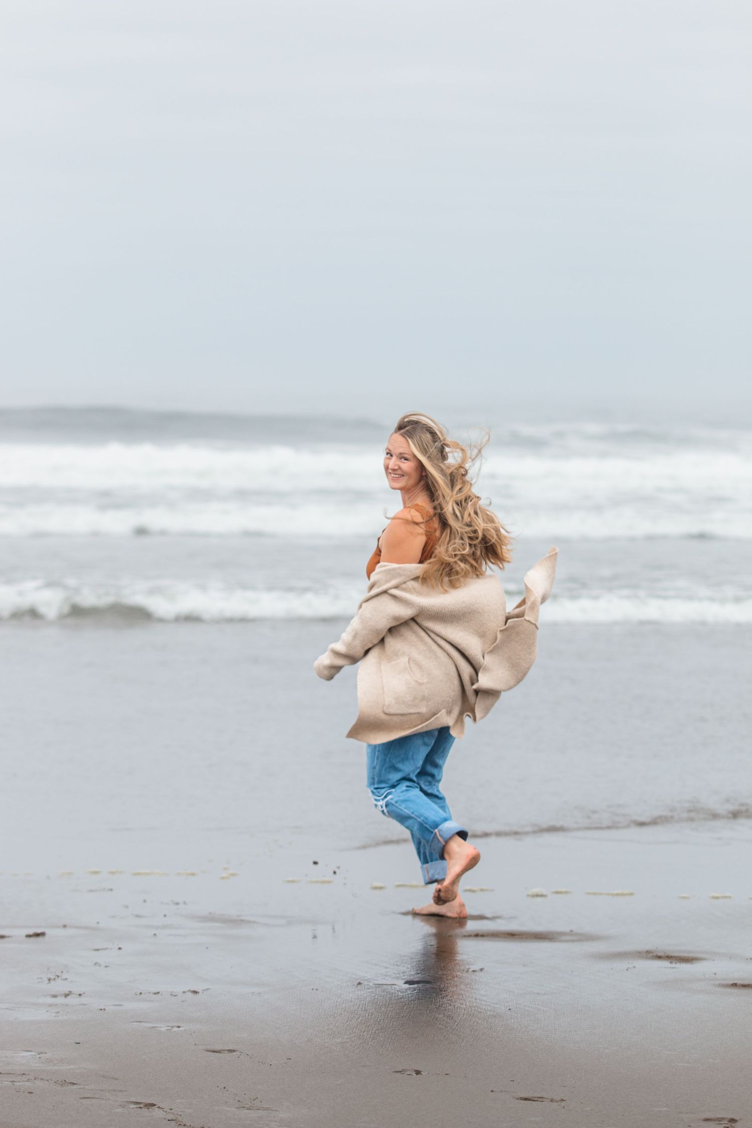 A person dances and twirls on a misty beach wearing an oversized beige sweater.