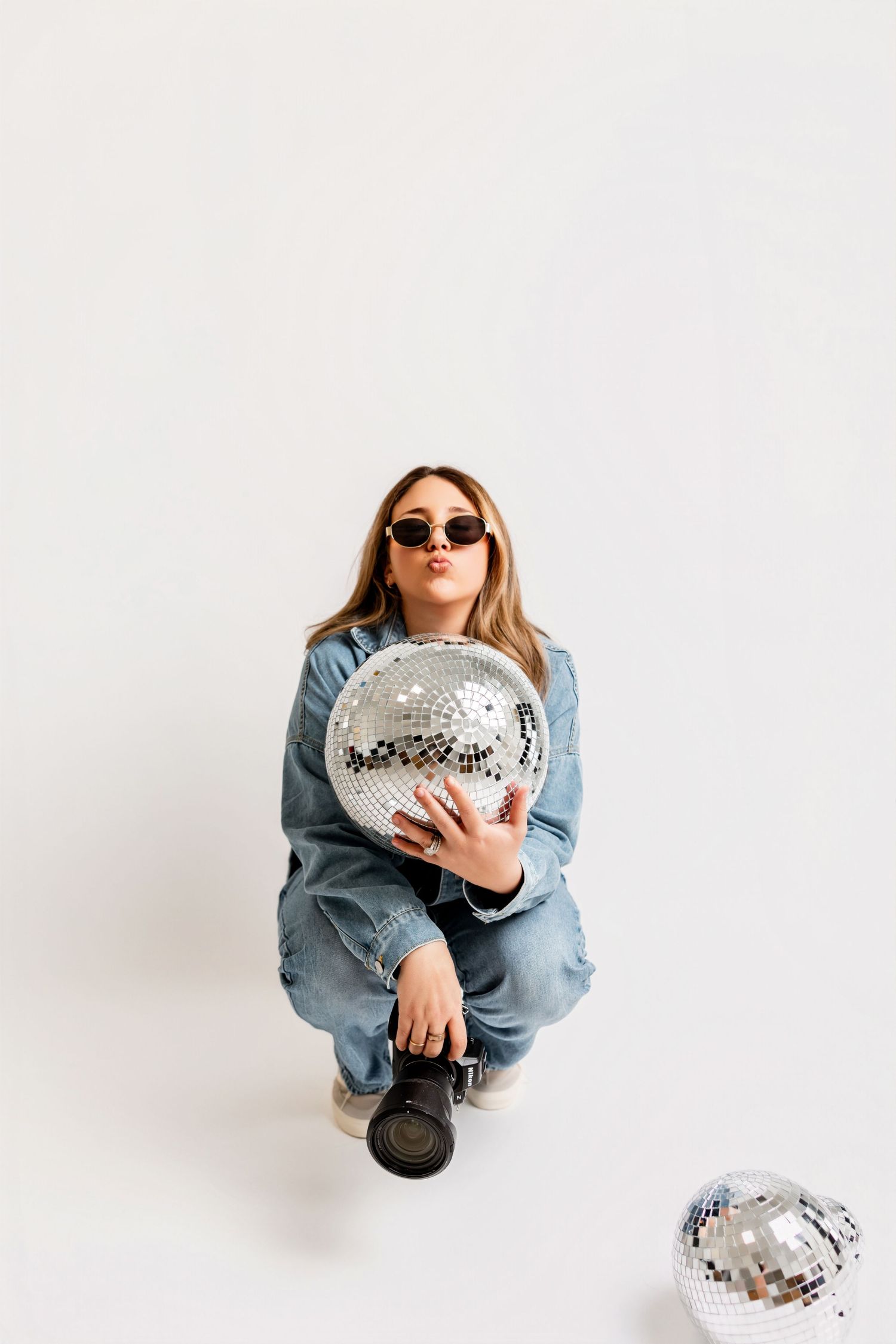Person in denim jacket and sunglasses poses with silver disco ball against white background in stylish photoshoot series.