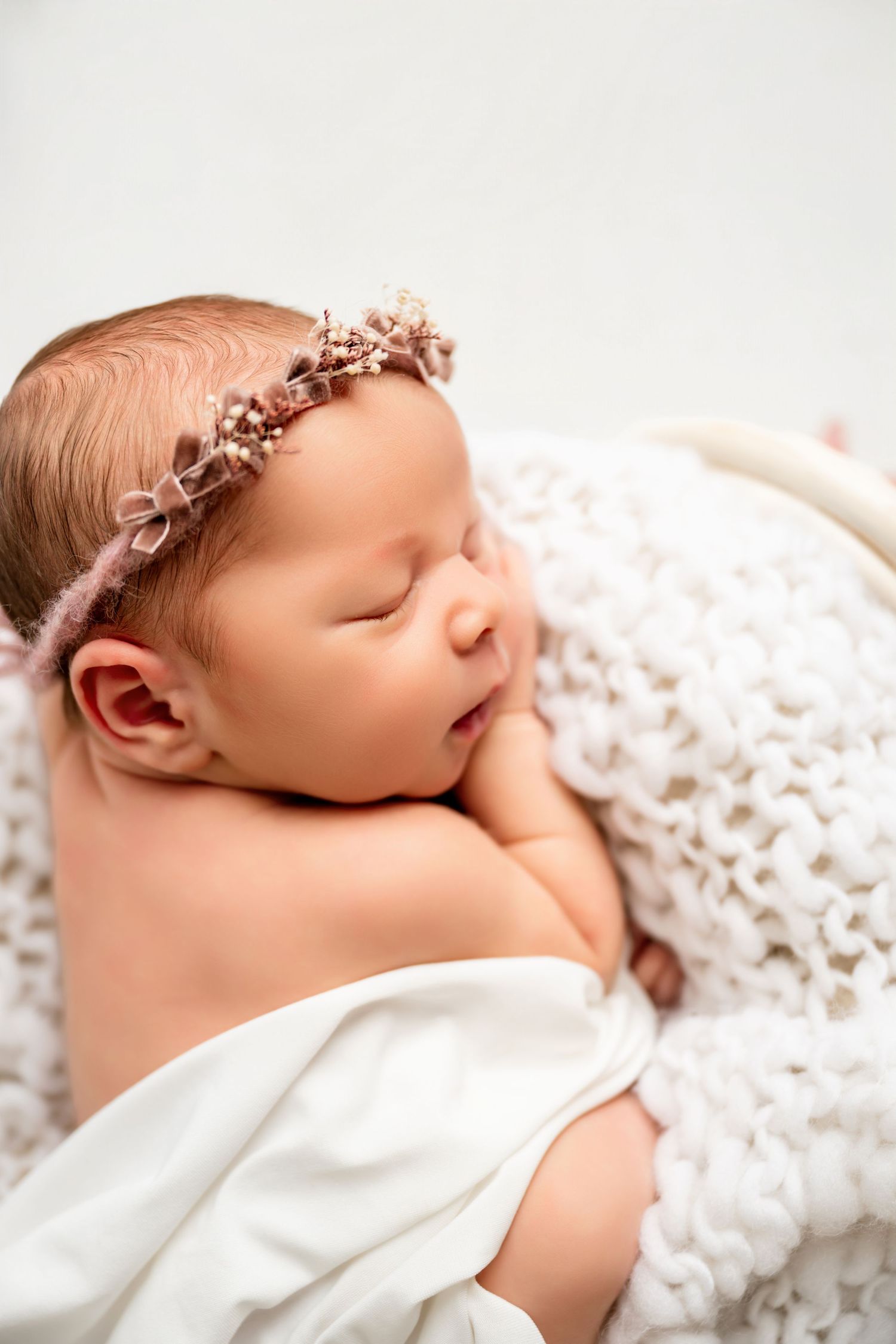 Sleeping newborn wearing delicate floral crown wrapped in white knit blanket.