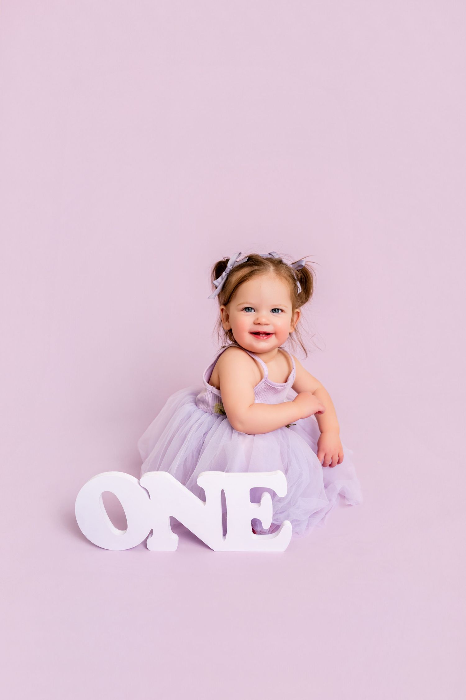 A baby in white dress lies on pink background with decorative white 'ONE' letters for first birthday photoshoot.