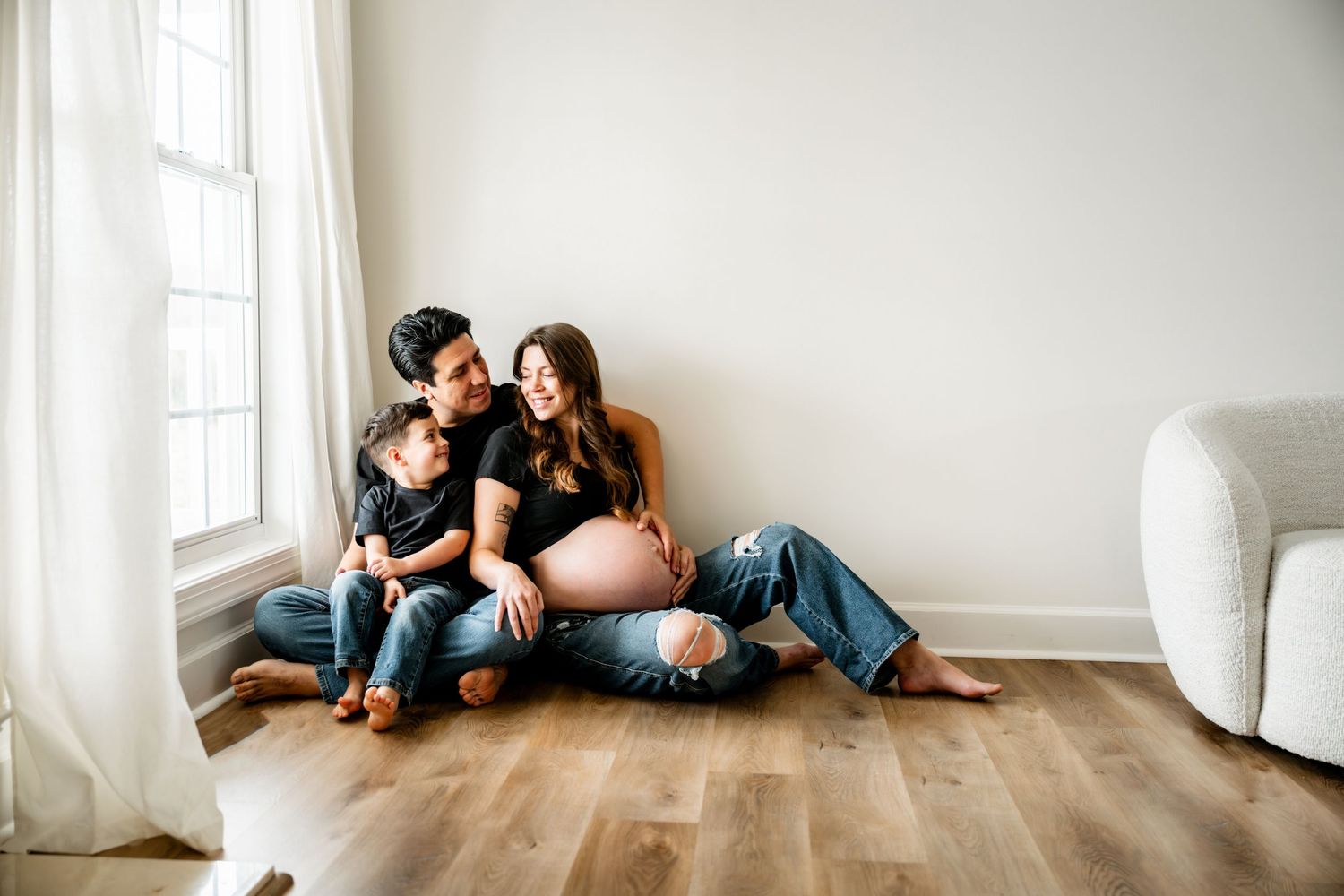 A family relaxes together on hardwood floors near a bright window and white curtains in a minimalist room.