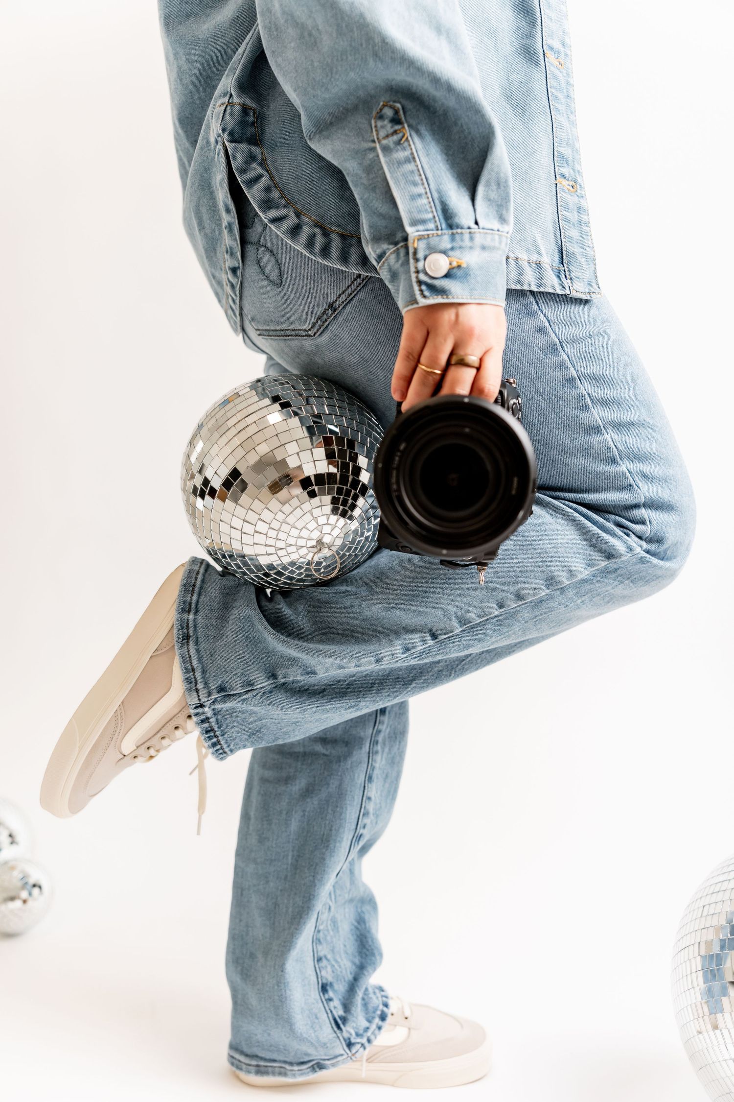 Person in light blue jeans holding a camera while posing with a disco ball on a white background.