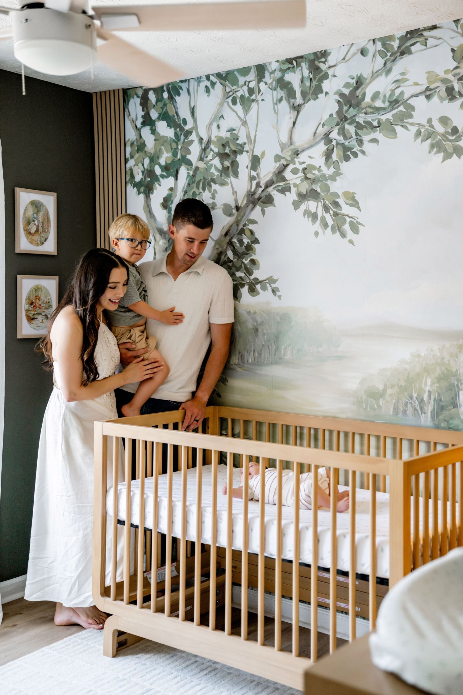Family of three admiring newborn baby in wooden crib, nursery with tree mural wall art.