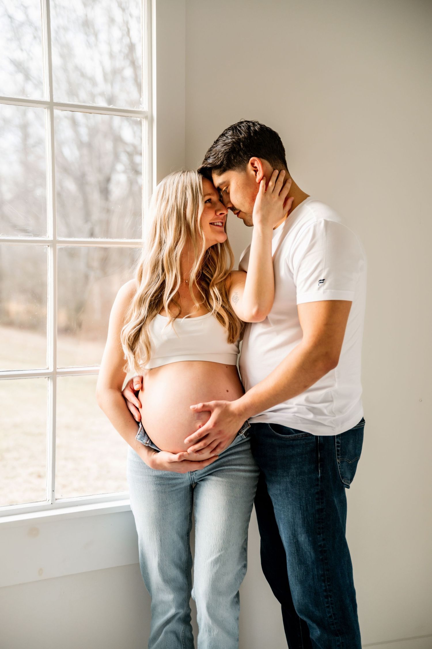 Expecting couple shares intimate moment by sunlit window as they lovingly embrace the pregnancy bump.