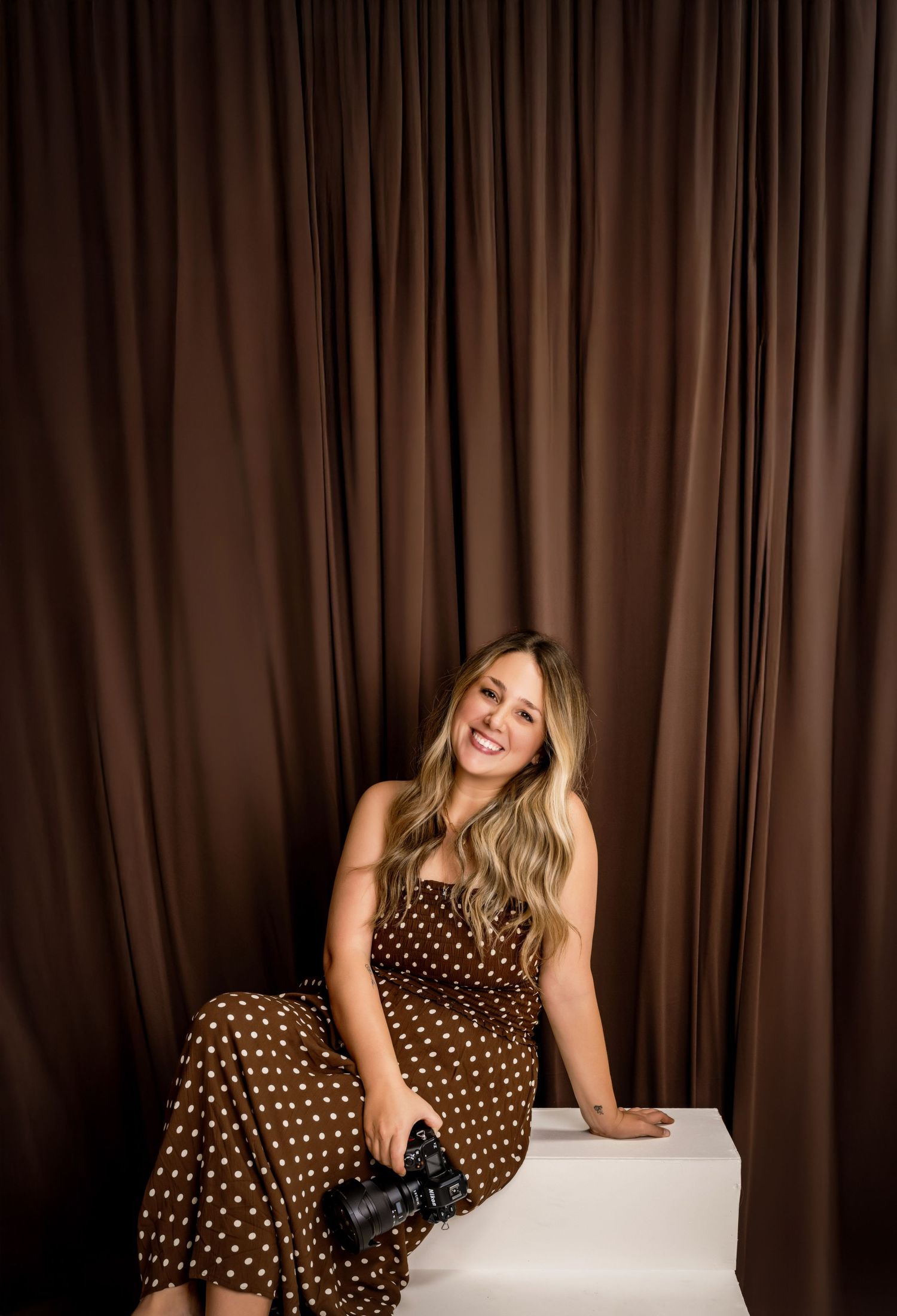 Woman in brown polka dot dress sits on white cube holding camera against brown draped backdrop.