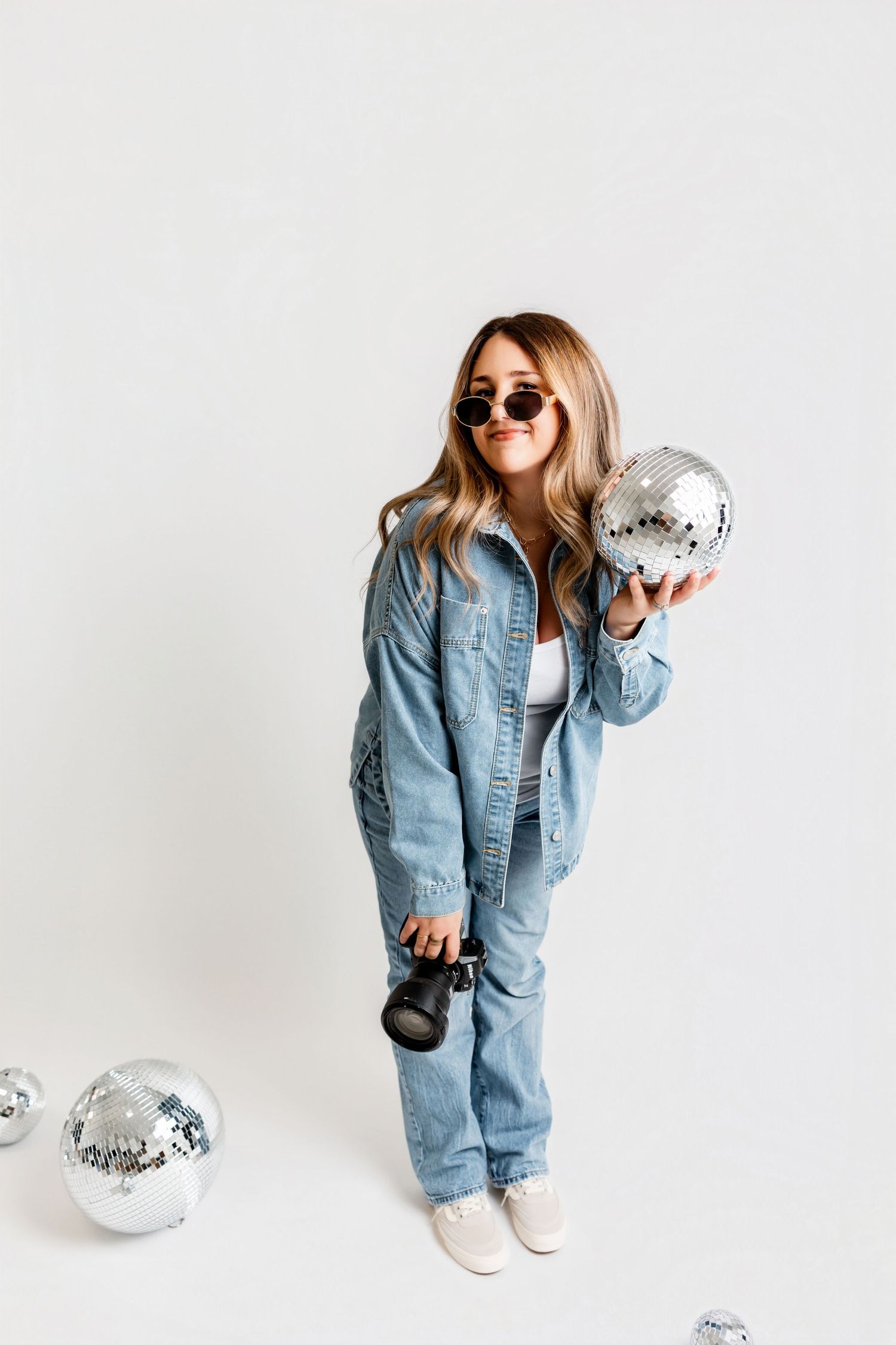 Playful fashion photo series of person in oversized denim jacket and sunglasses posing with disco balls against white backdrop.