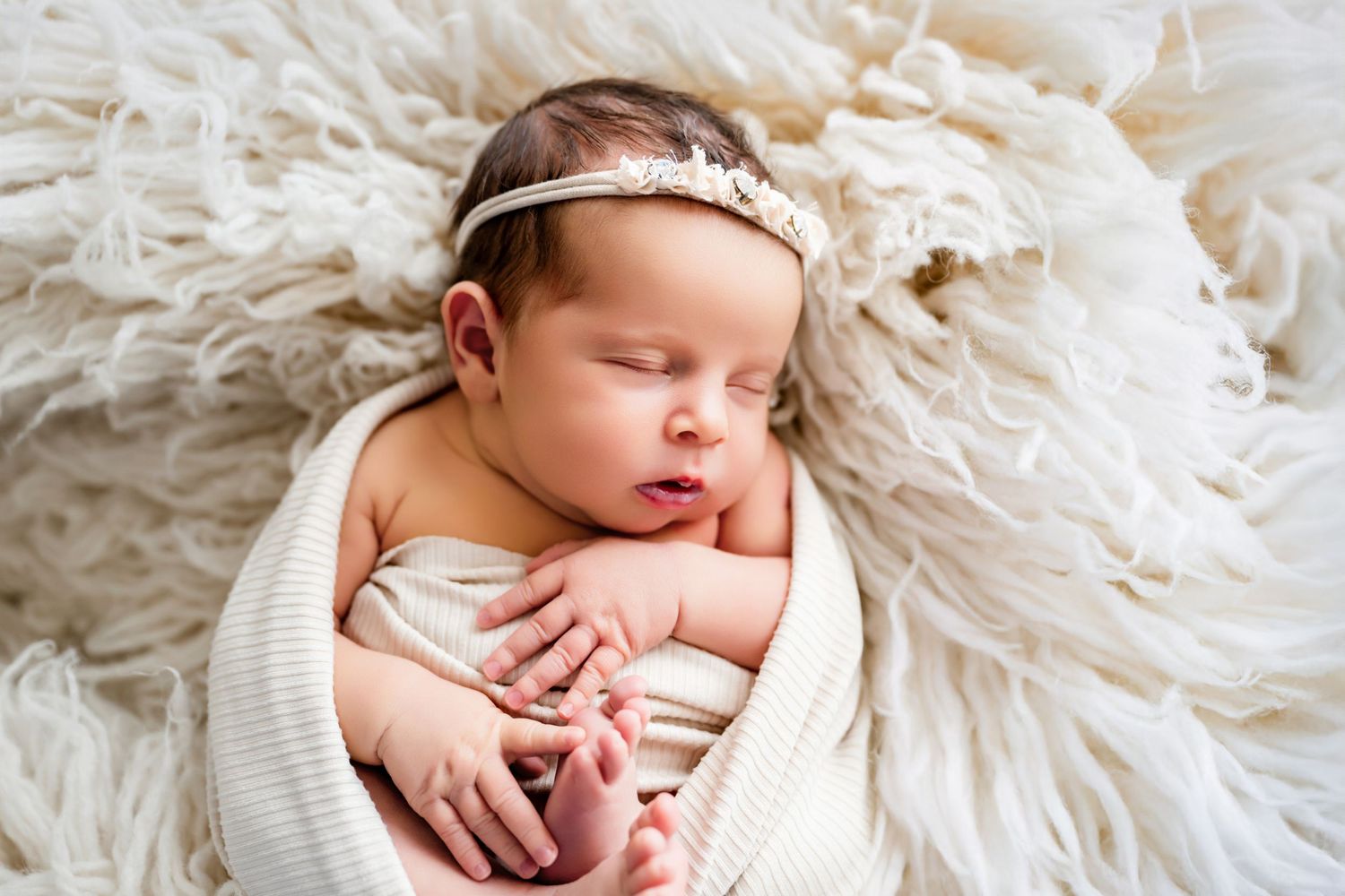 A sleeping newborn baby swaddled in soft white fabric lies peacefully on a textured cream blanket.