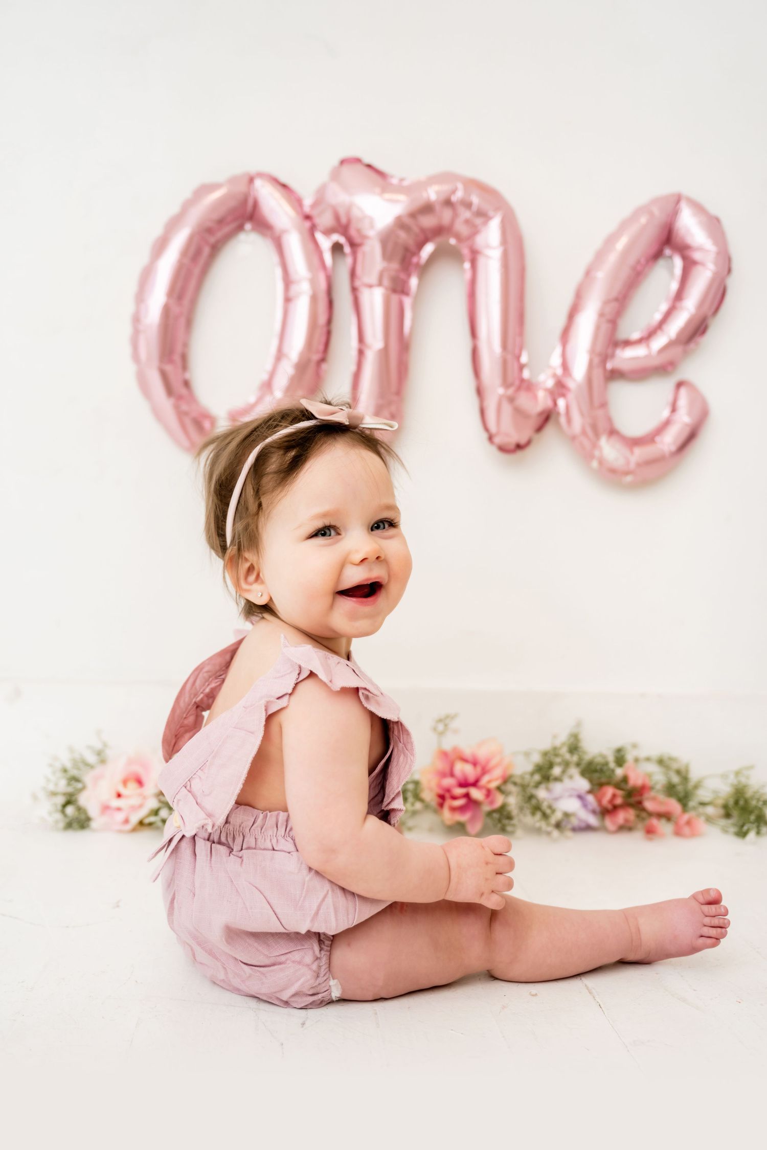 Baby in pink romper sitting under rose gold ONE balloon decoration.