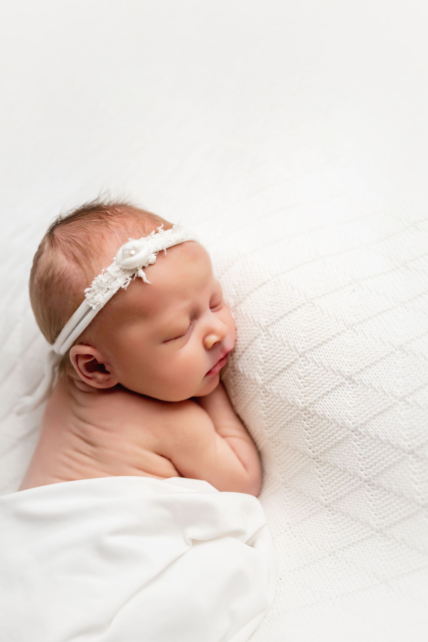 Sleeping newborn baby wearing white headband wrapped in soft white blanket against light background.