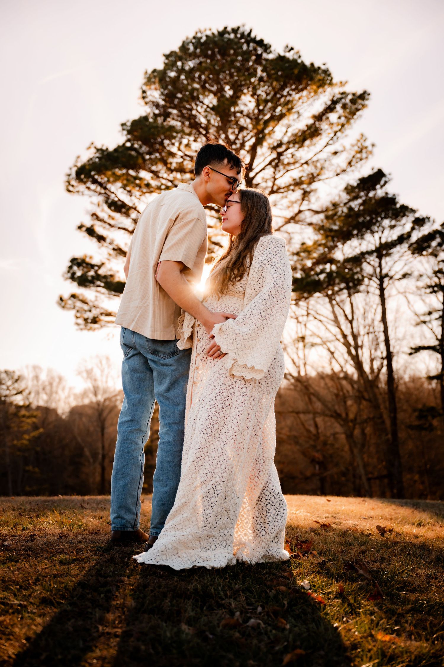A couple embraces in a romantic sunset moment beneath a large pine tree in a golden-lit field.