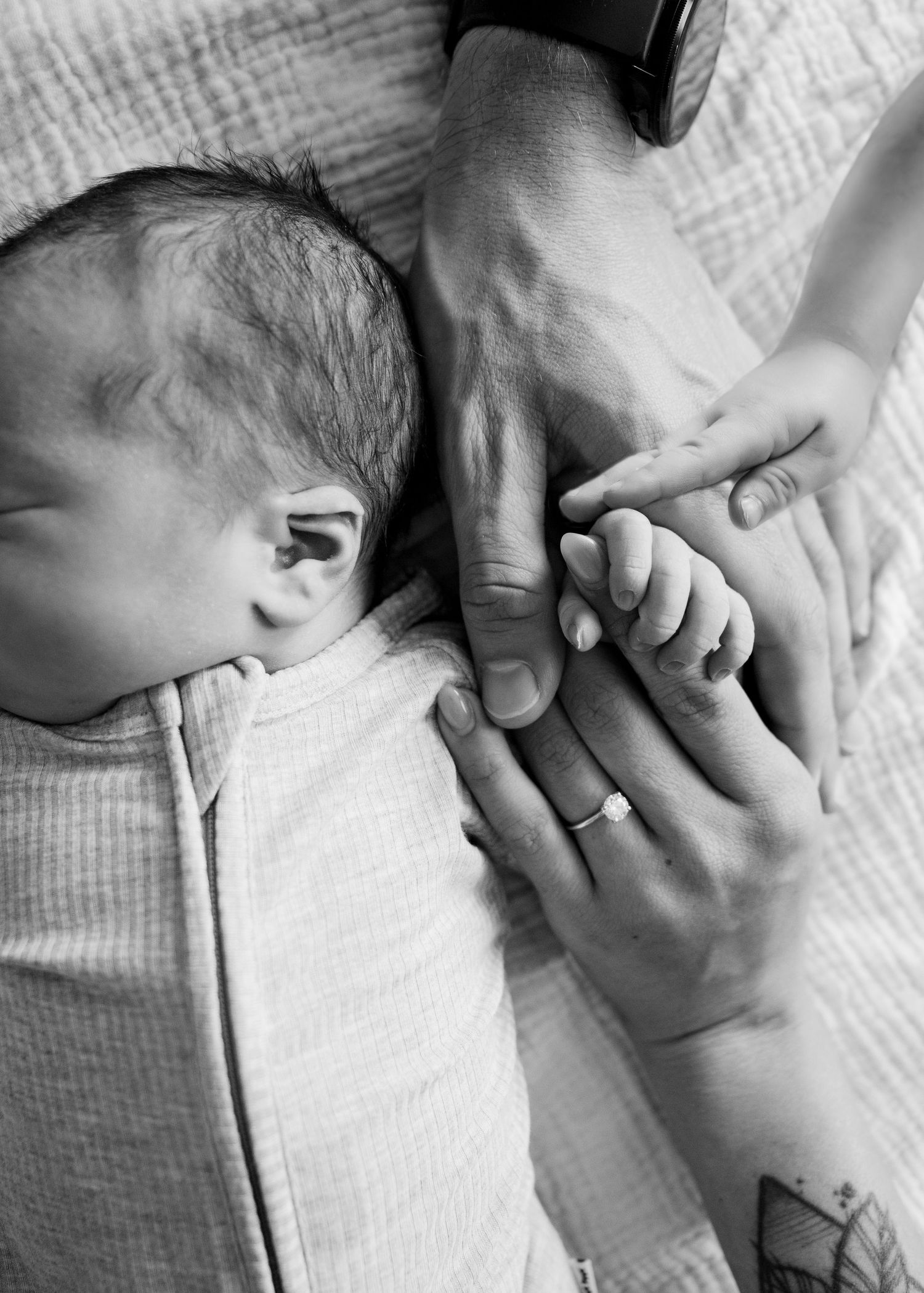 Black and white image of tiny baby hand holding adult fingers on textured blanket.
