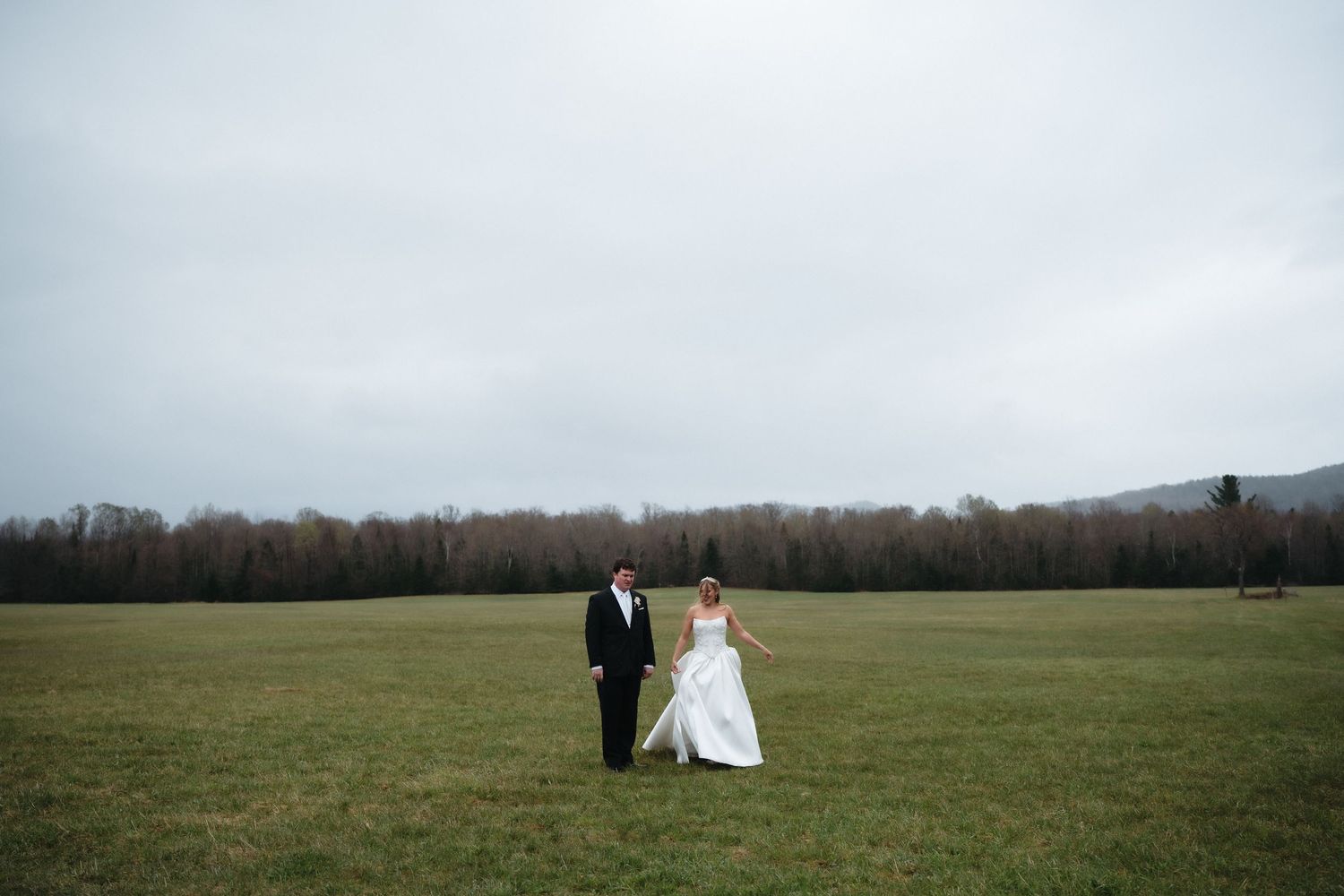 Bride and groom walking hand in hand across a lush green field on a cloudy day at an outdoor wedding venue.