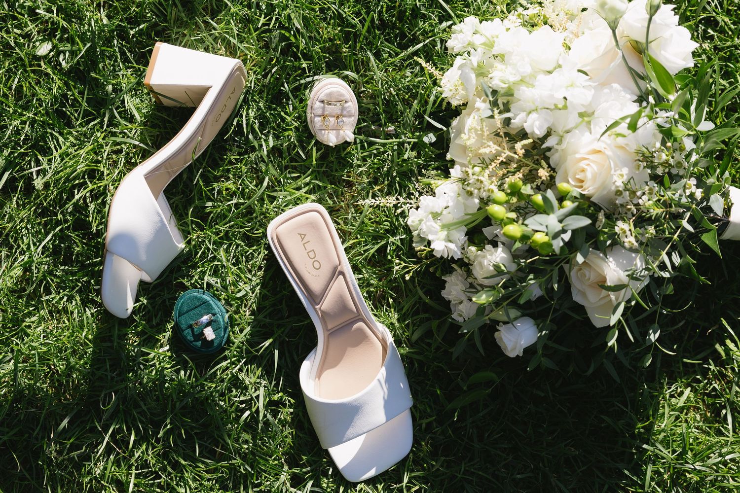 White bridal heels, bouquet of white flowers, and elegant jewelry accessories arranged on lush green grass.