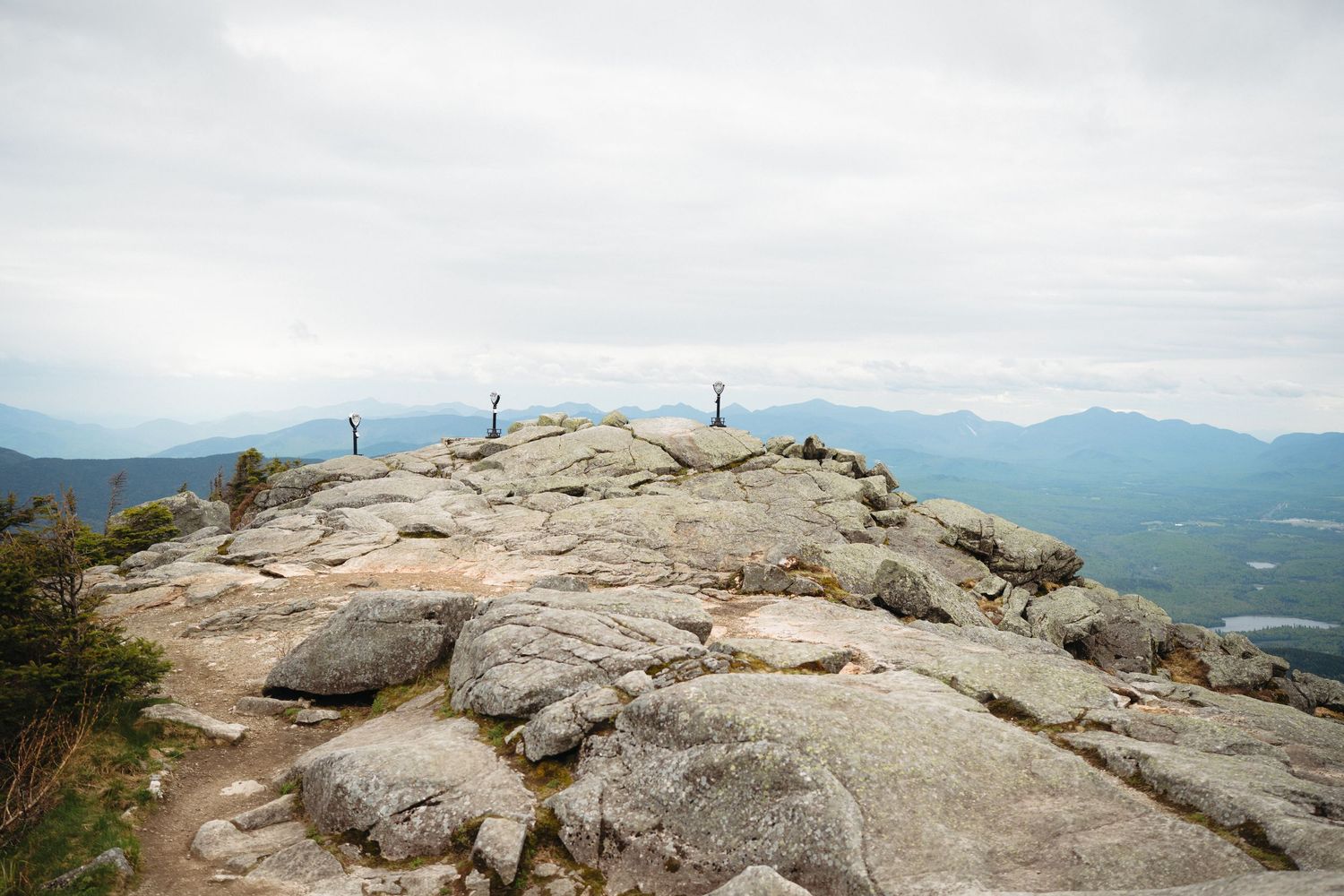 Rocky mountain summit with hikers and panoramic views of misty blue mountain ranges under an overcast sky.