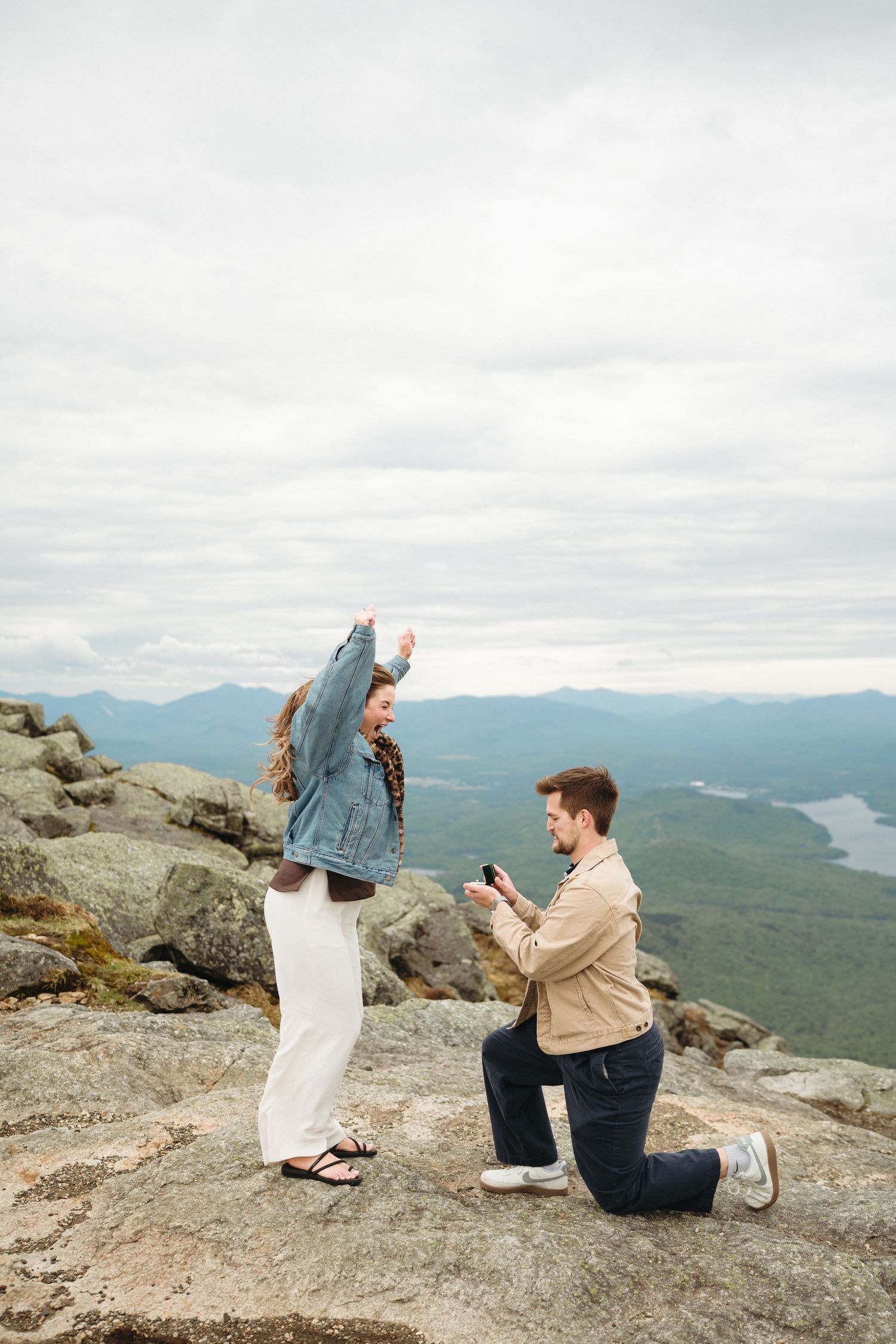 Man proposes on one knee atop rocky mountain summit, woman raises arms in joy, scenic valley and lake backdrop.