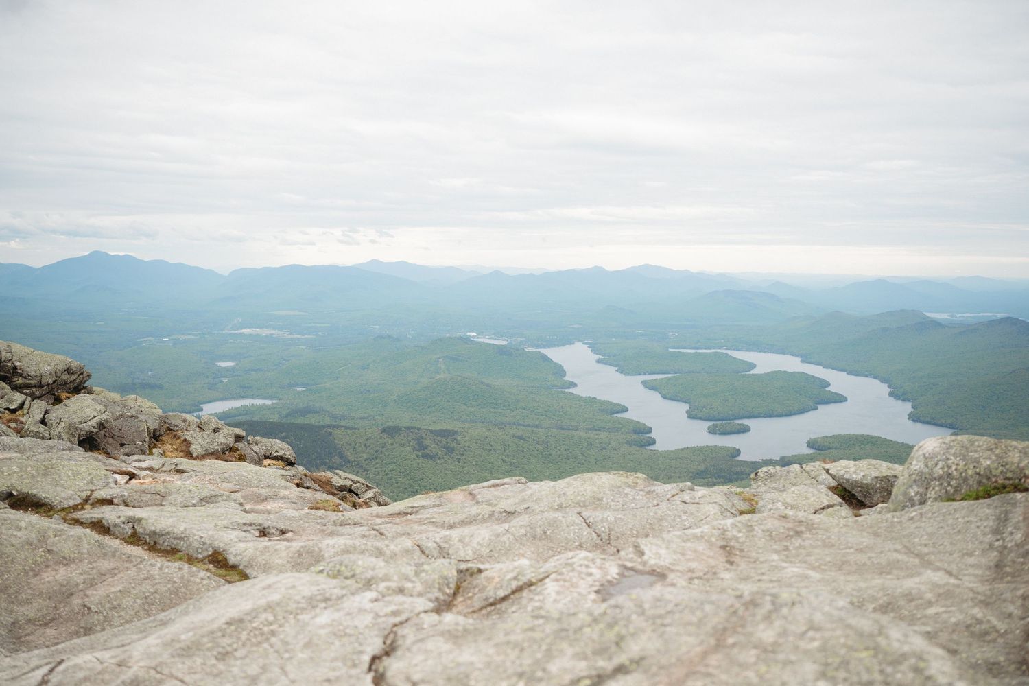 Aerial view from rocky mountain summit overlooking lush green forest and winding lake under overcast sky.