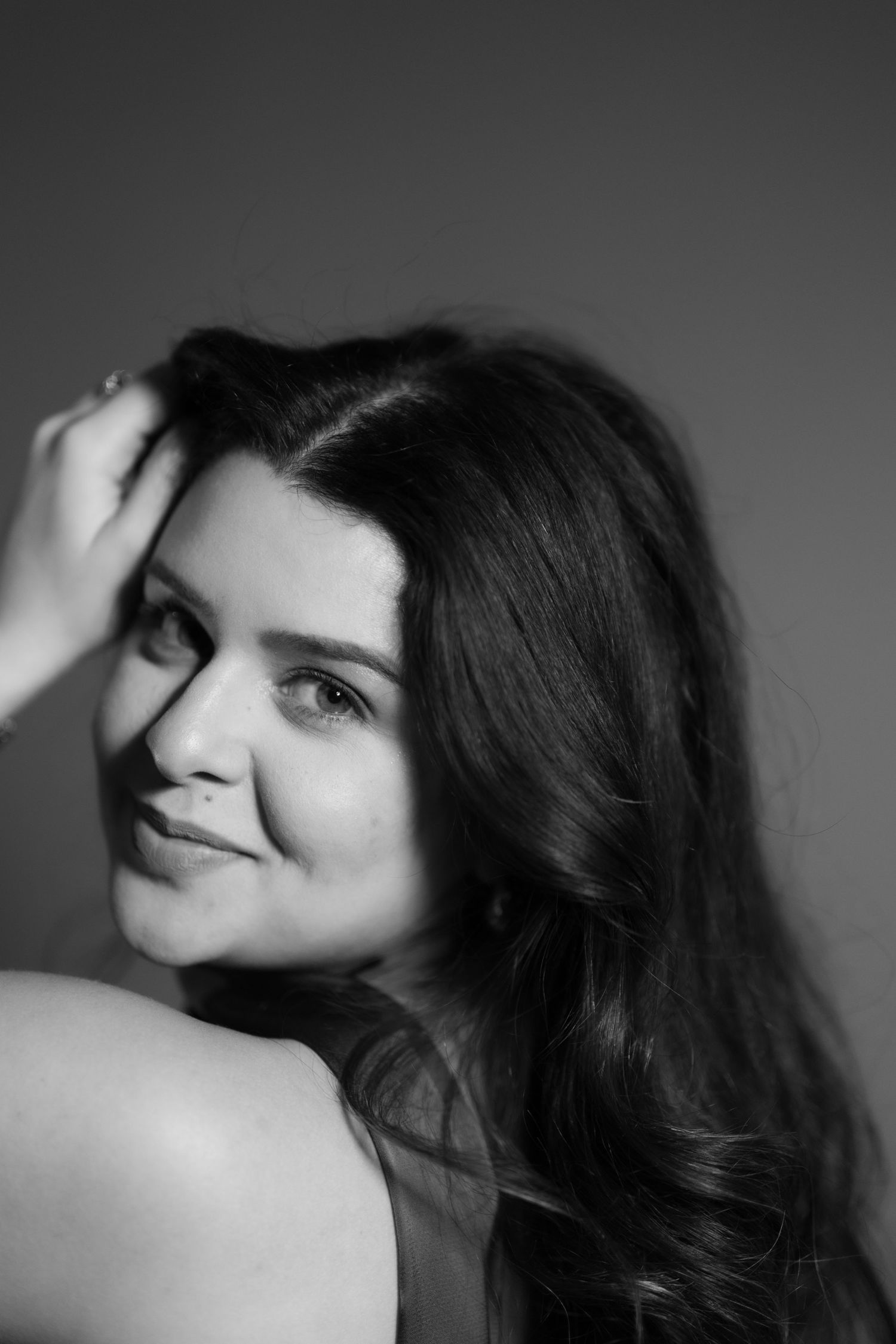 Black and white portrait of a young woman with long dark hair, smiling softly while touching her hair.