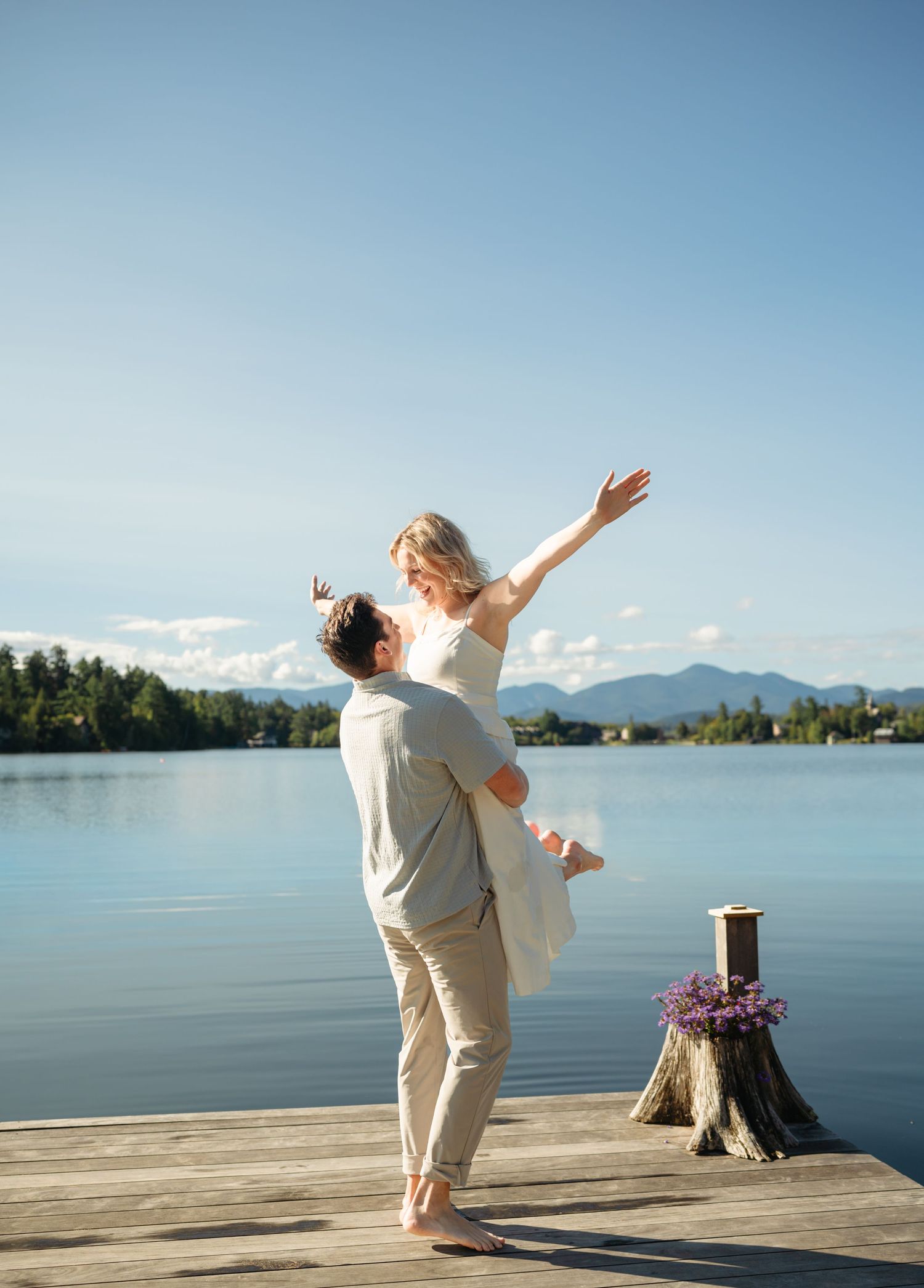 A groom lifts his bride on a lakeside dock, mountains in the background, on a sunny wedding day.