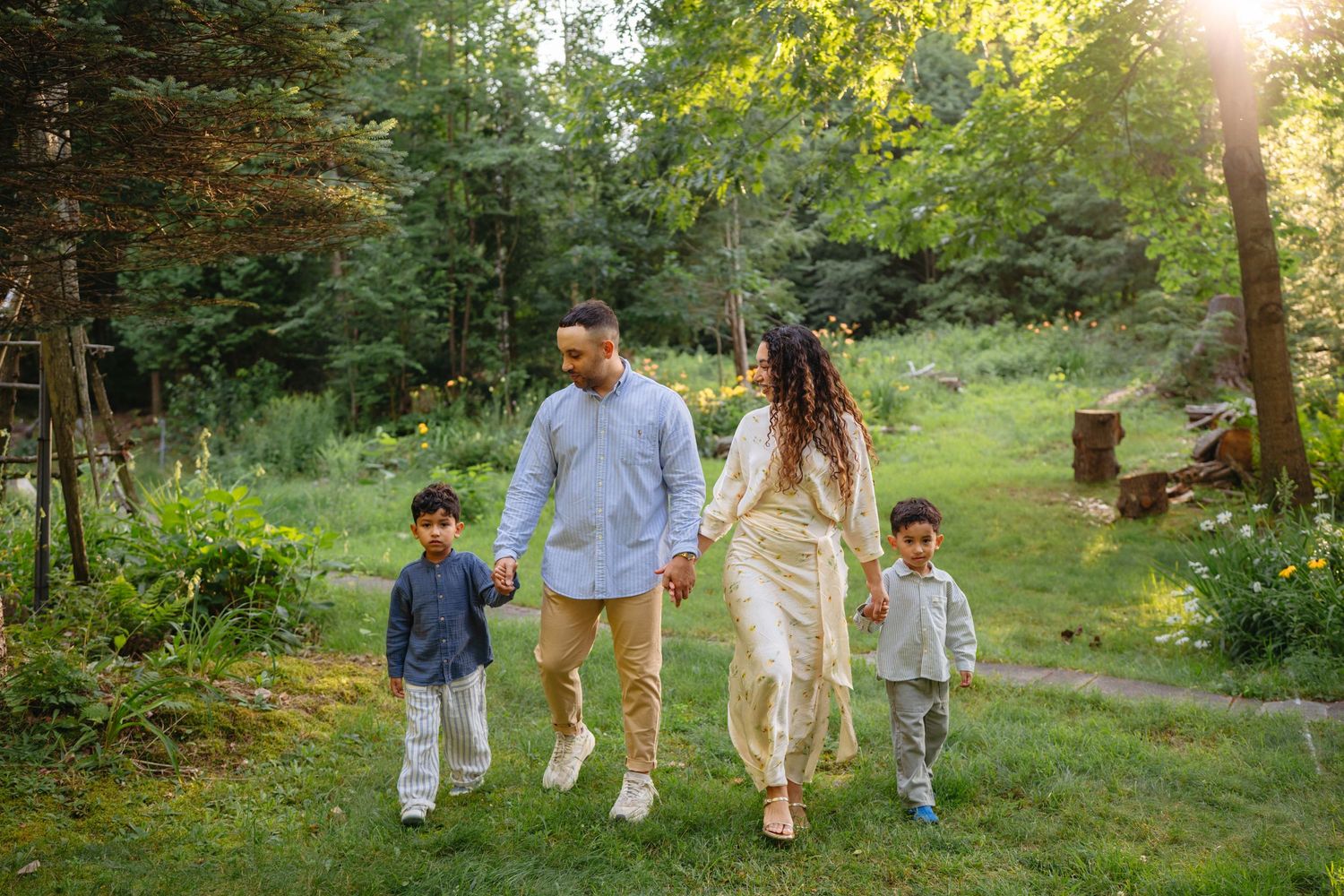 Family of four walking hand-in-hand through a lush green forest path during golden hour sunset.