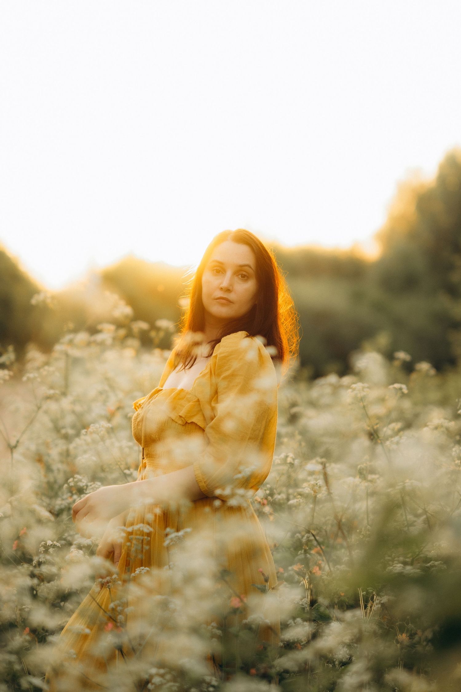 a woman in a field of cow parsley, wearing a yellow dress looking into the camera
