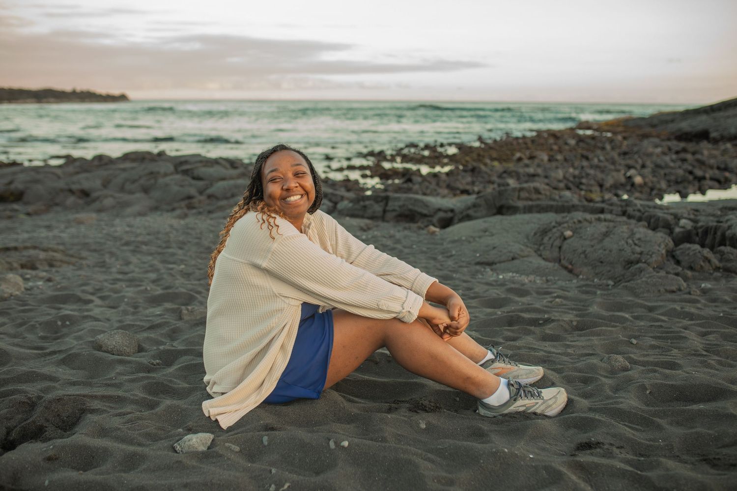 Young woman smiling and sitting on rocky beach shore with turquoise ocean waves in background.