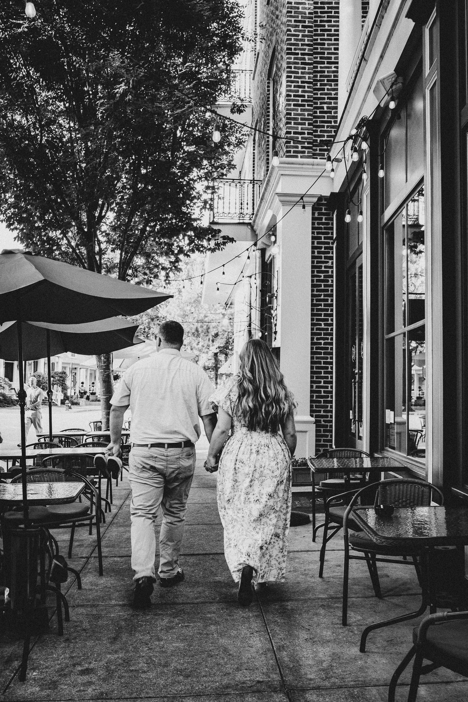 A couple walking on a sidewalk past outdoor cafe seating on a sunny day in black and white.
