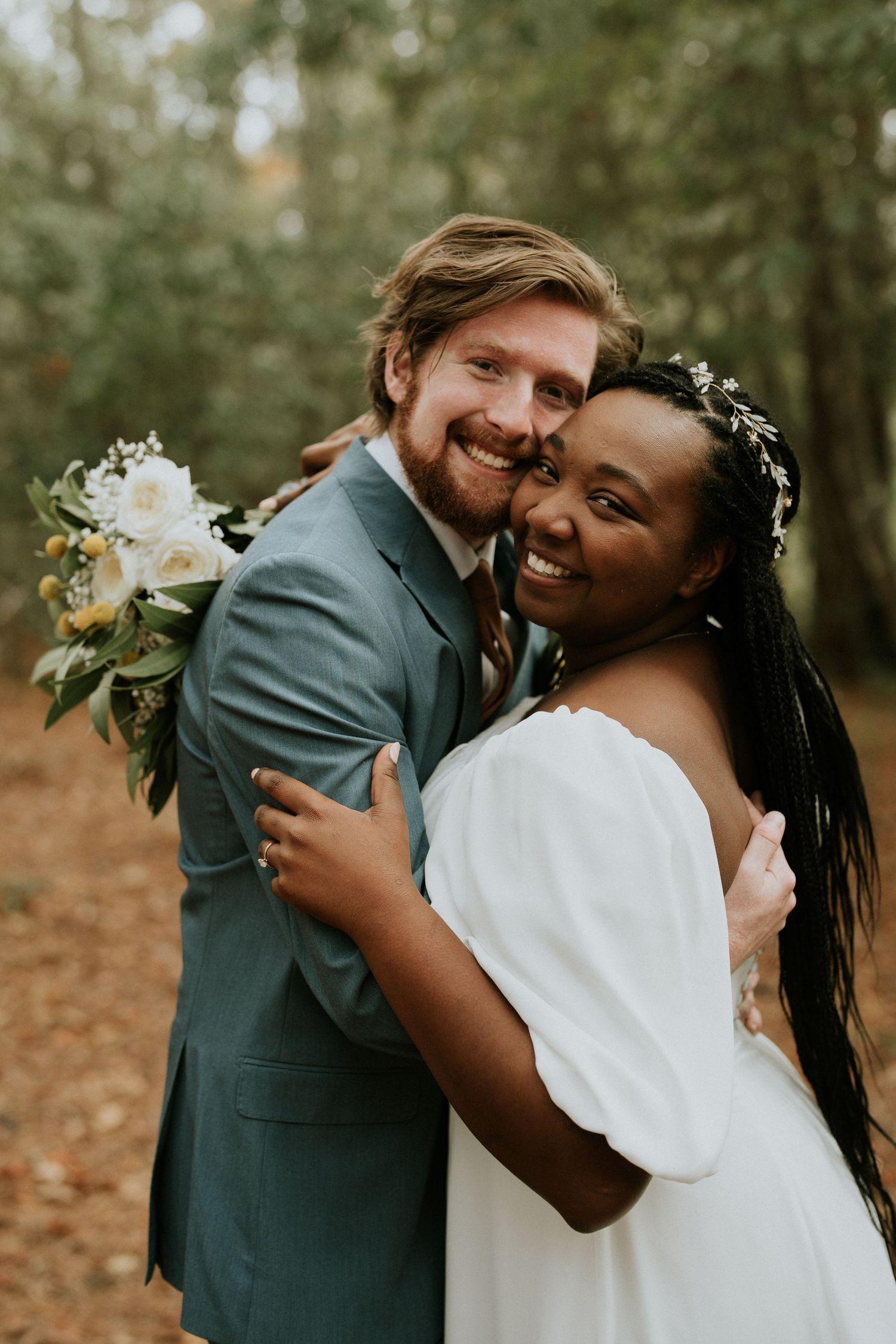 Newlyweds embrace and smile in forest setting with white flowers.