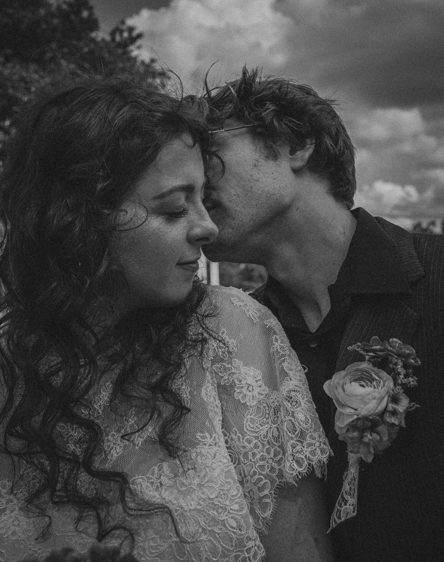 Black and white wedding photo of groom kissing bride on cheek, bride in lace dress with curly hair outdoors.
