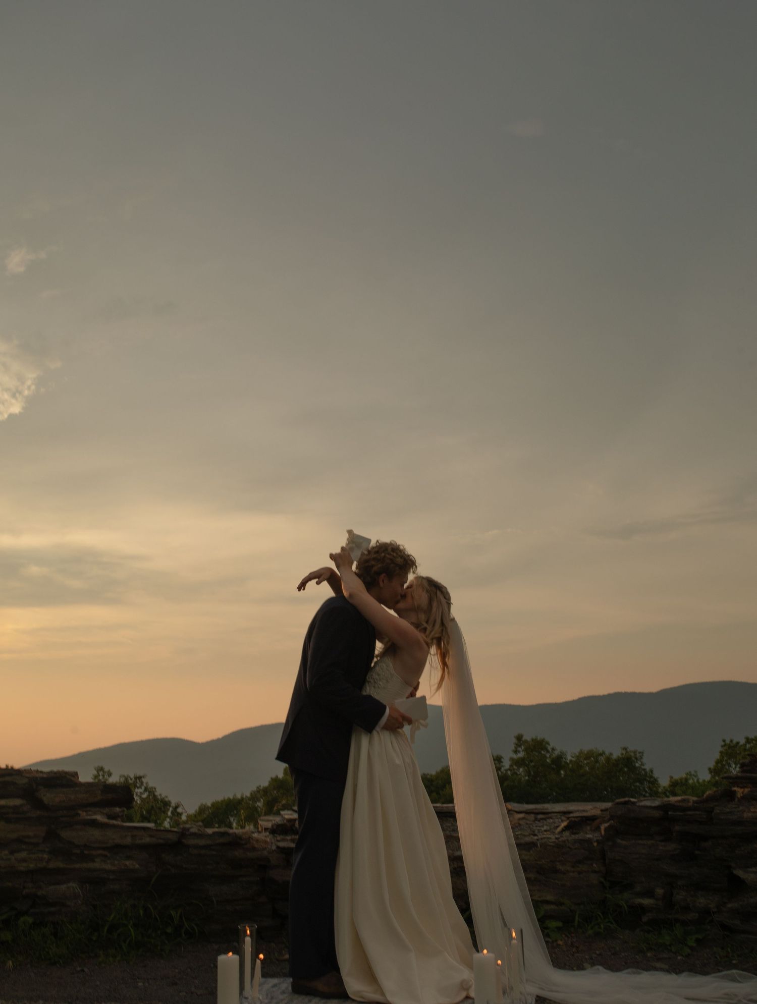 Bride and groom share a kiss at sunset with mountain views and candles, during an outdoor wedding ceremony.