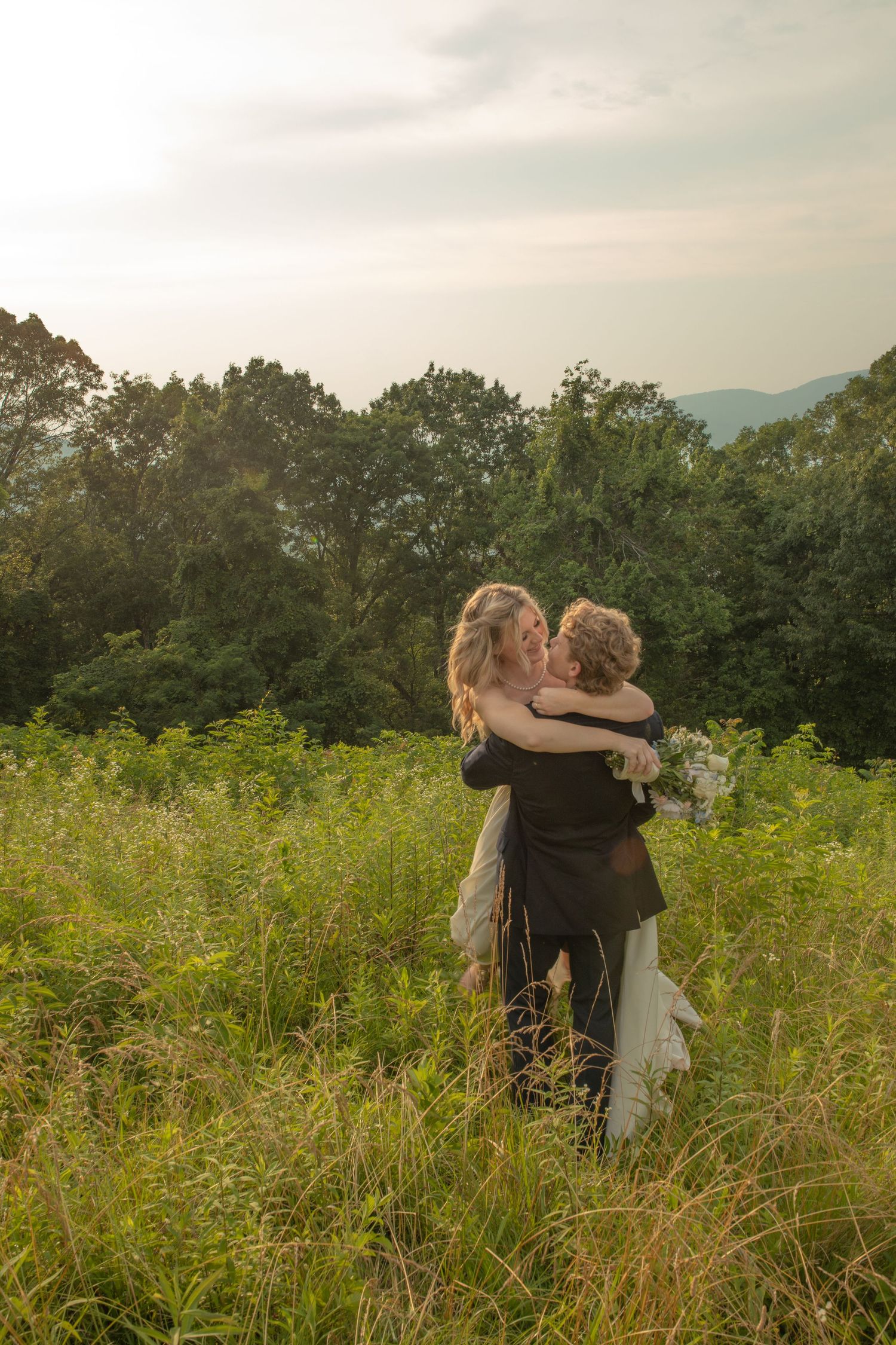 Couple embracing and kissing in a lush green meadow, wearing wedding attire, with trees and mountains in the background.