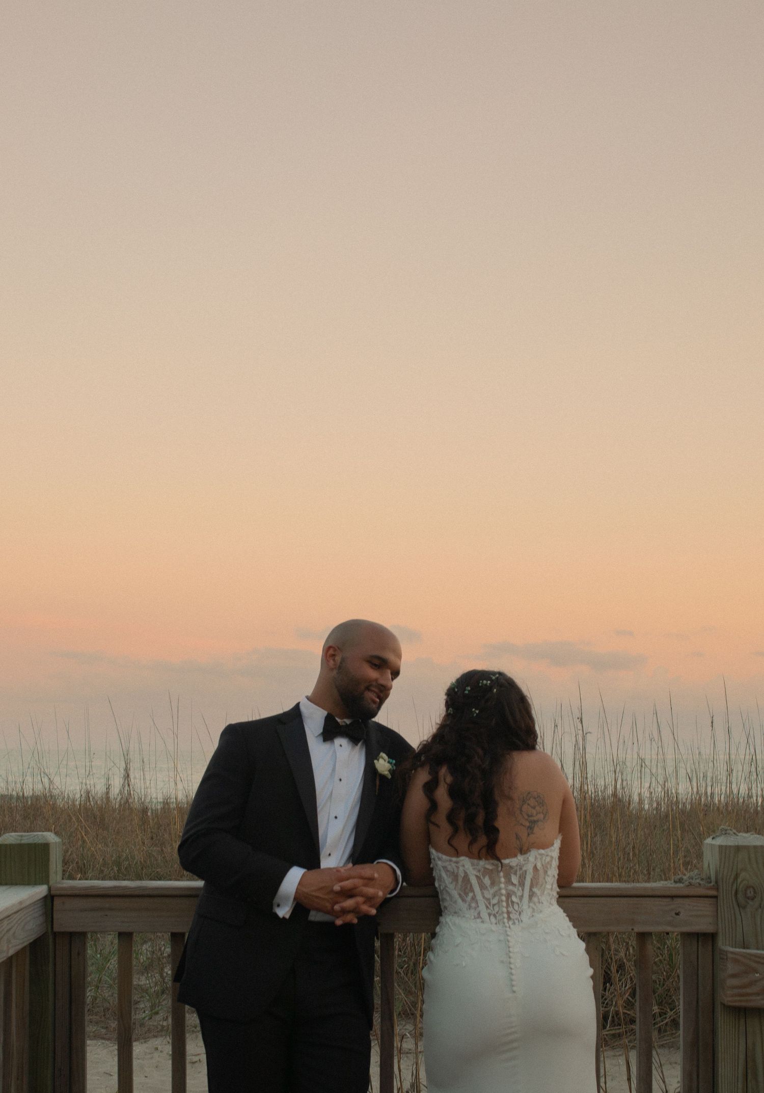 A couple in wedding attire shares a romantic moment on a beach deck at sunset, the bride in a lace gown and groom in a tuxedo.