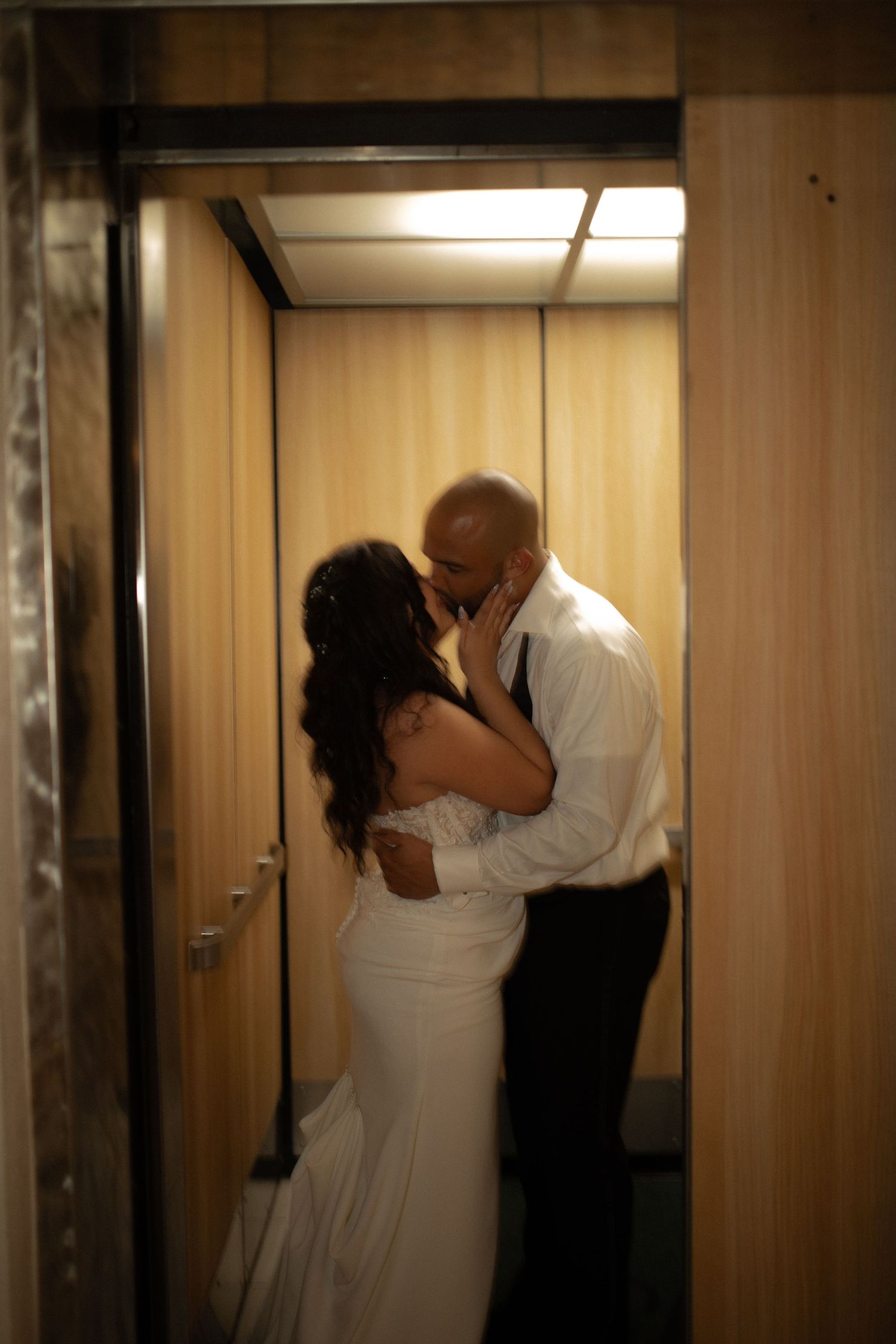 Bride and groom sharing a romantic kiss inside a warmly lit elevator on their wedding day.