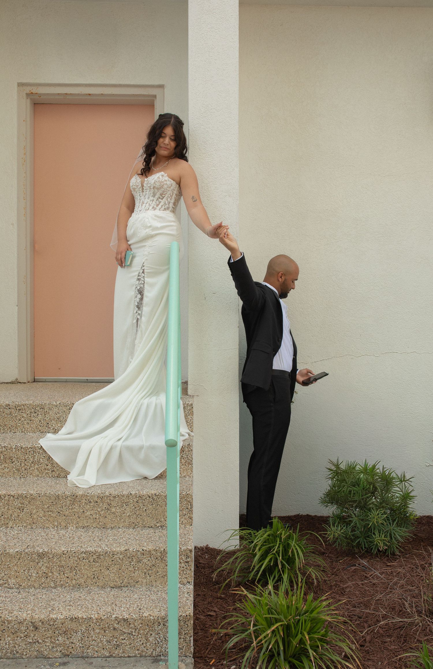 Bride in white gown stands on steps holding groom's hand in black tuxedo outside a pastel building.
