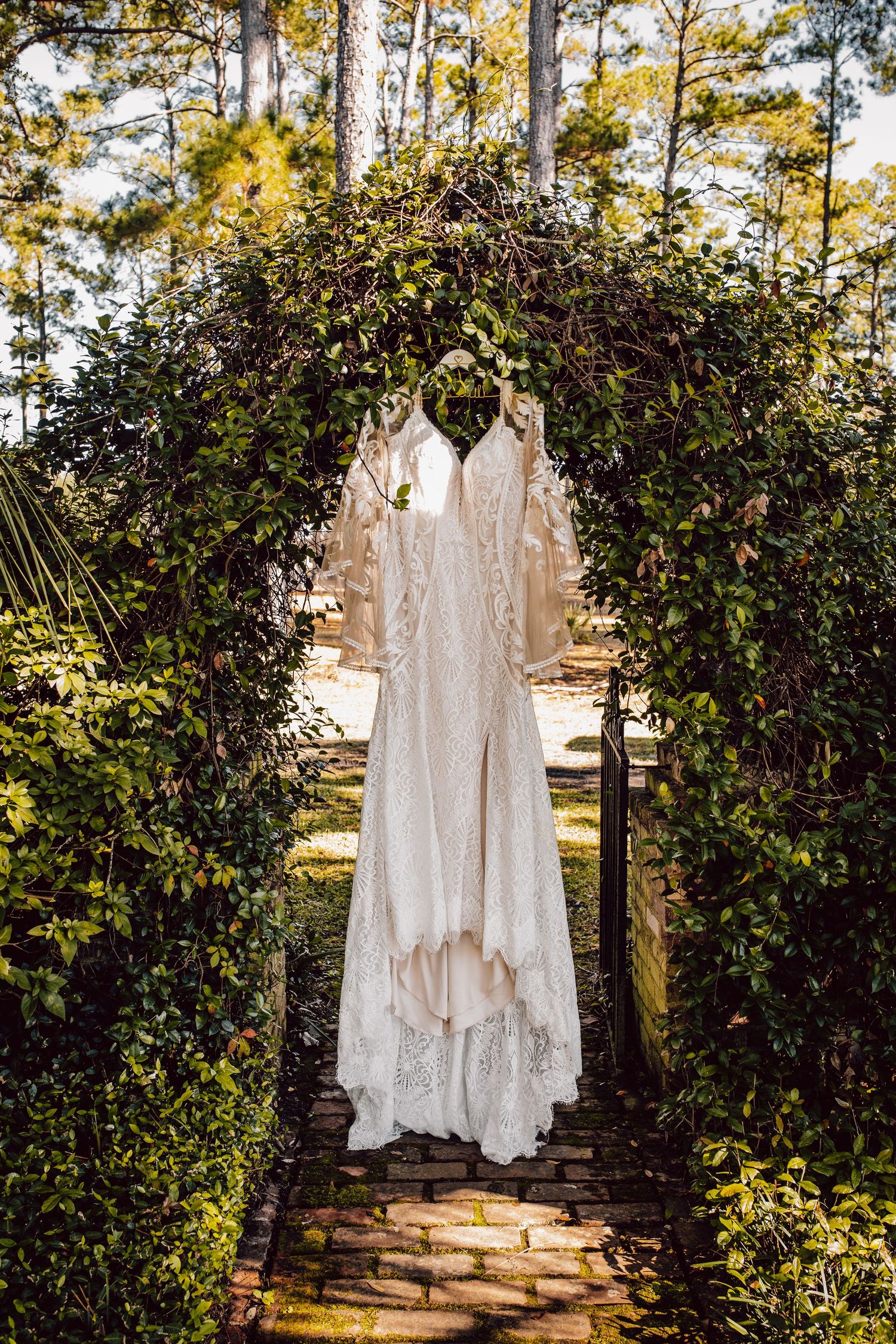 A beautiful white lace wedding dress hangs under a floral garden arch with greenery in natural outdoor lighting.
