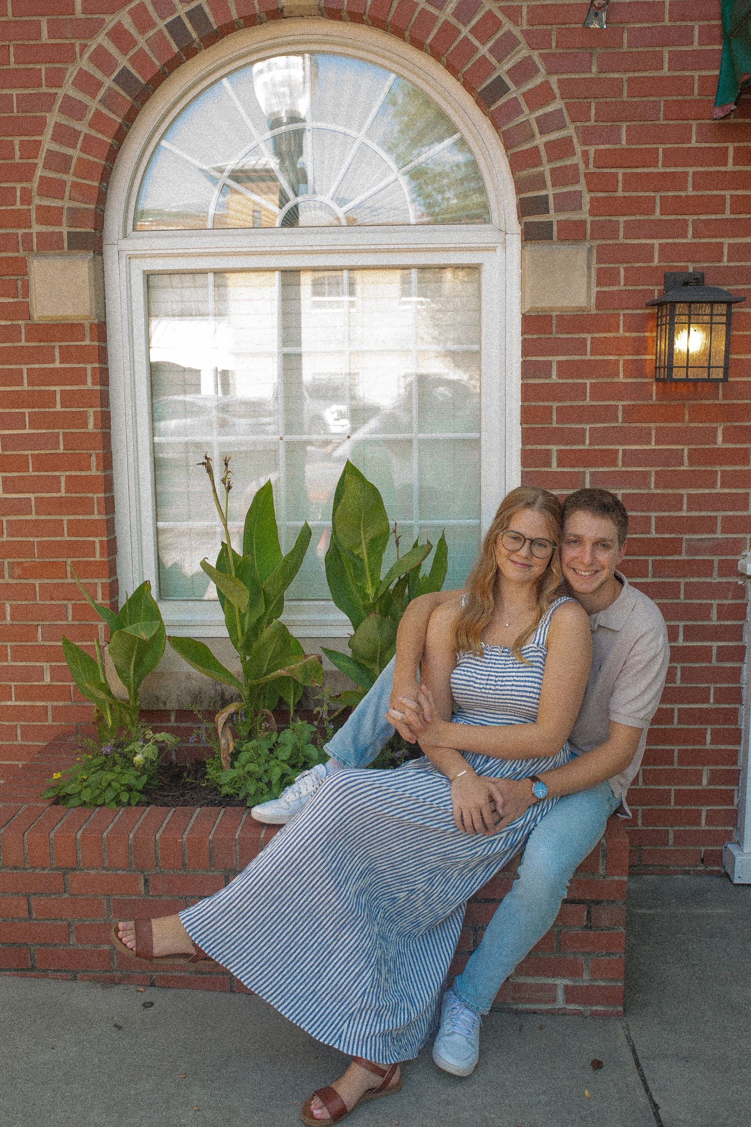 A couple sits together on a brick porch ledge near an arched window and lush green plants.