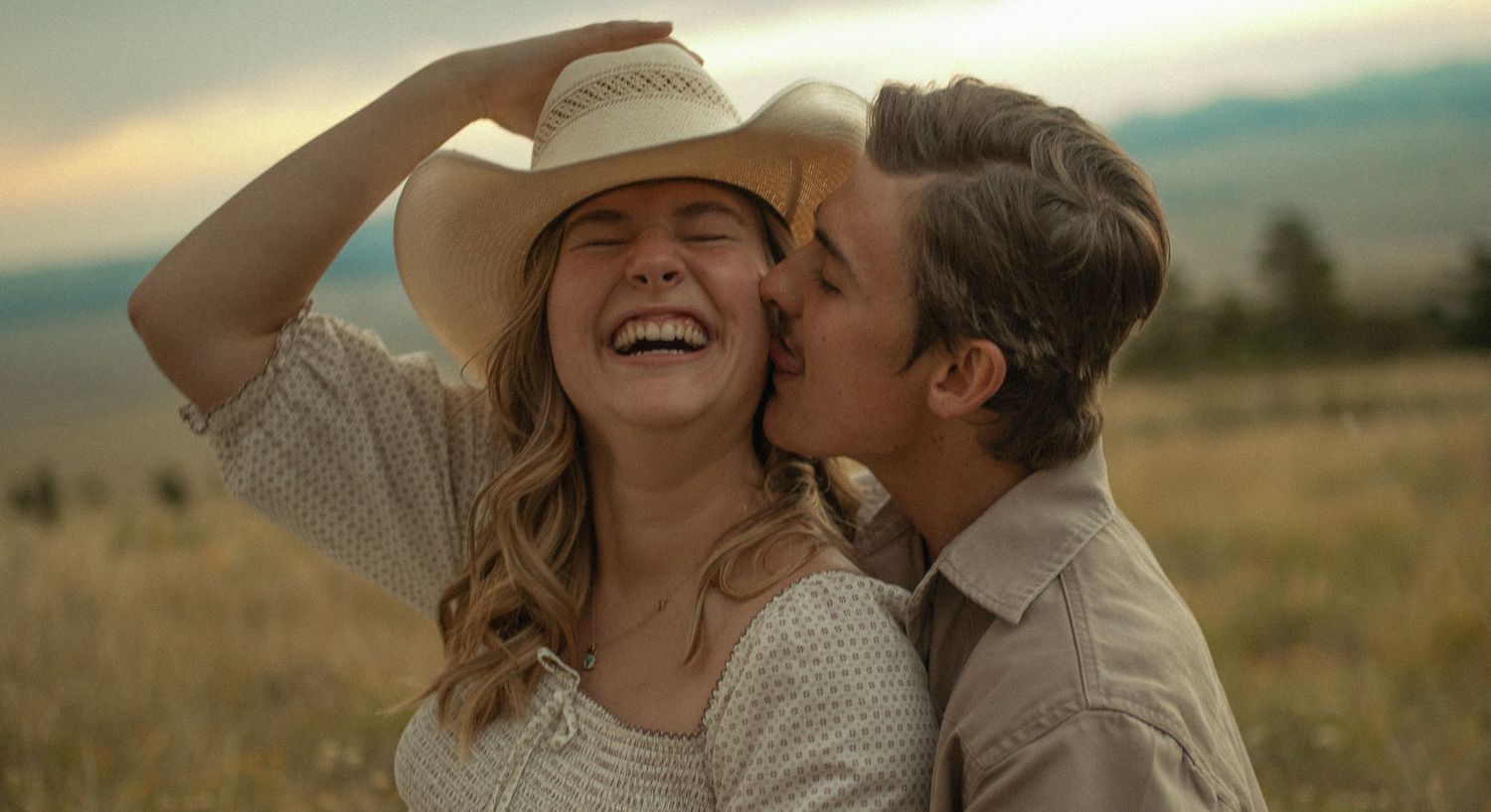 Happy couple in a field, woman in cowboy hat laughing as man kisses her cheek at golden hour.