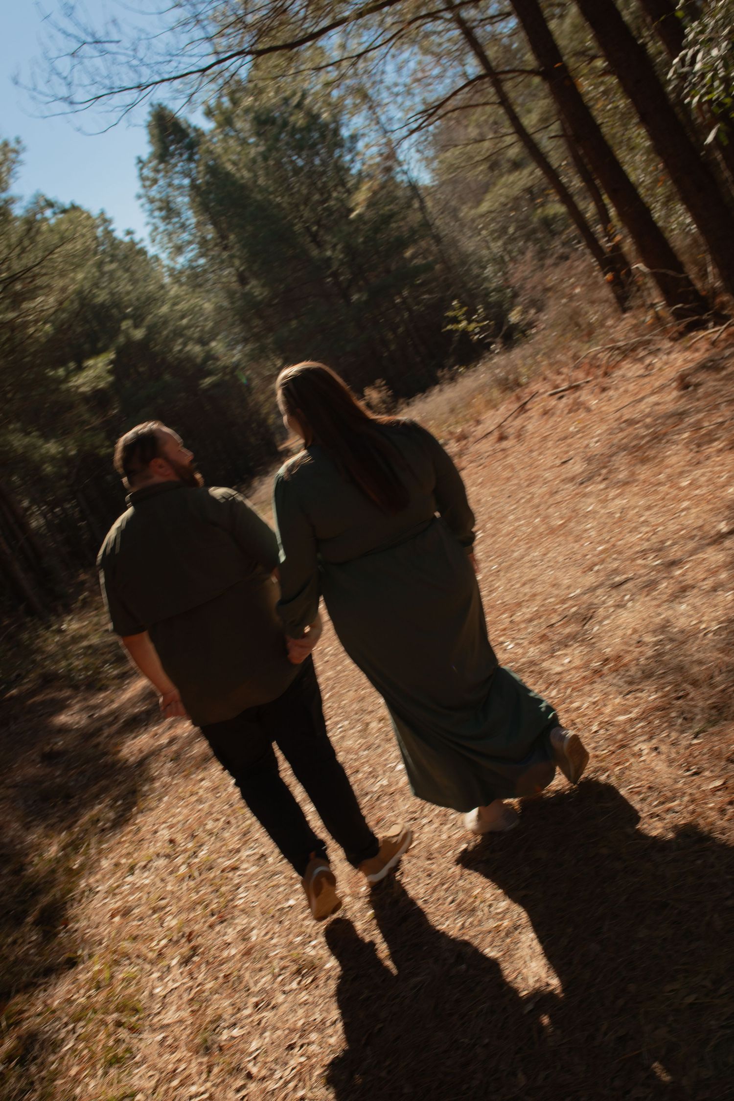 Two silhouettes walk together on a dirt path through a wooded area during sunset creating long shadows.