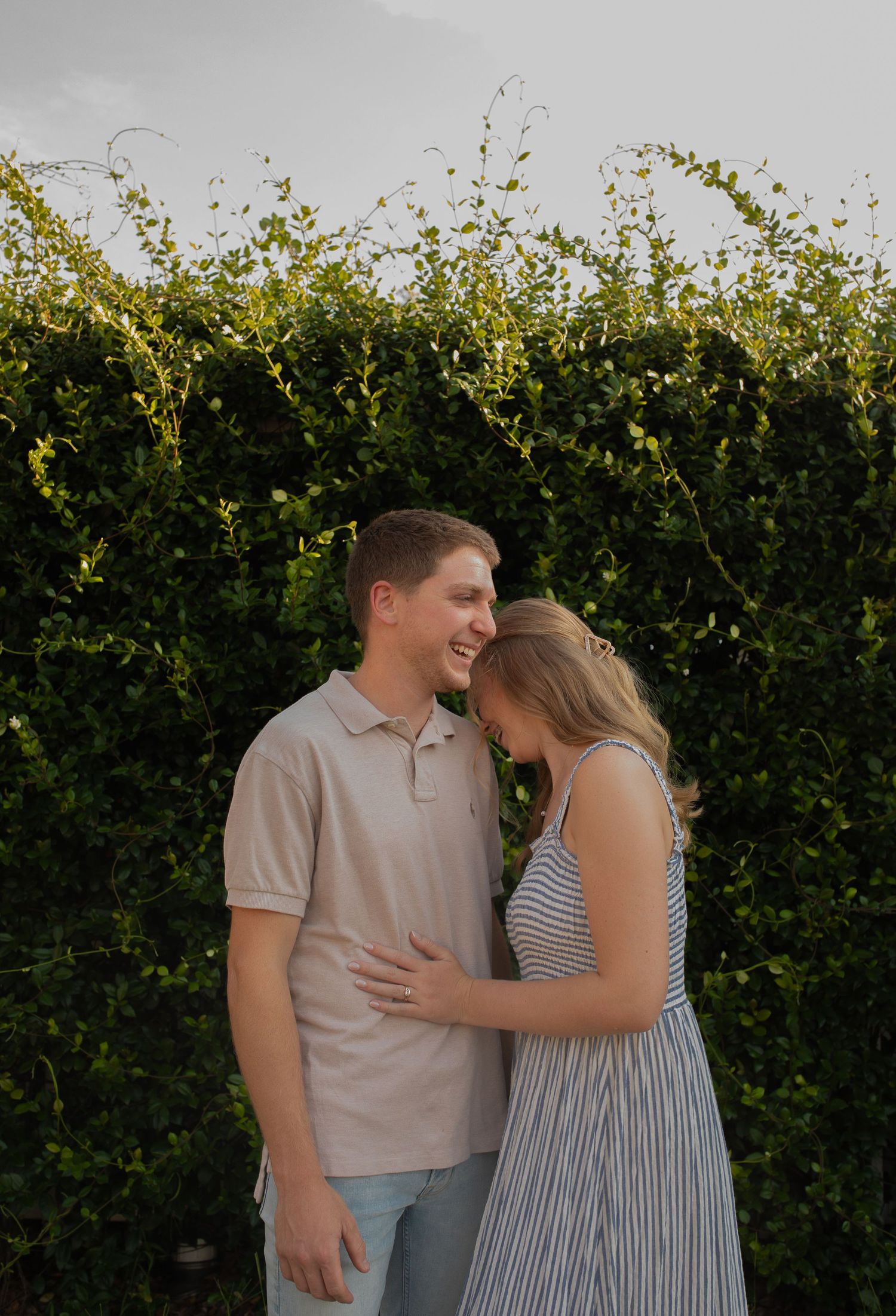 A couple shares a tender moment outdoors in front of lush green shrubbery at sunset.