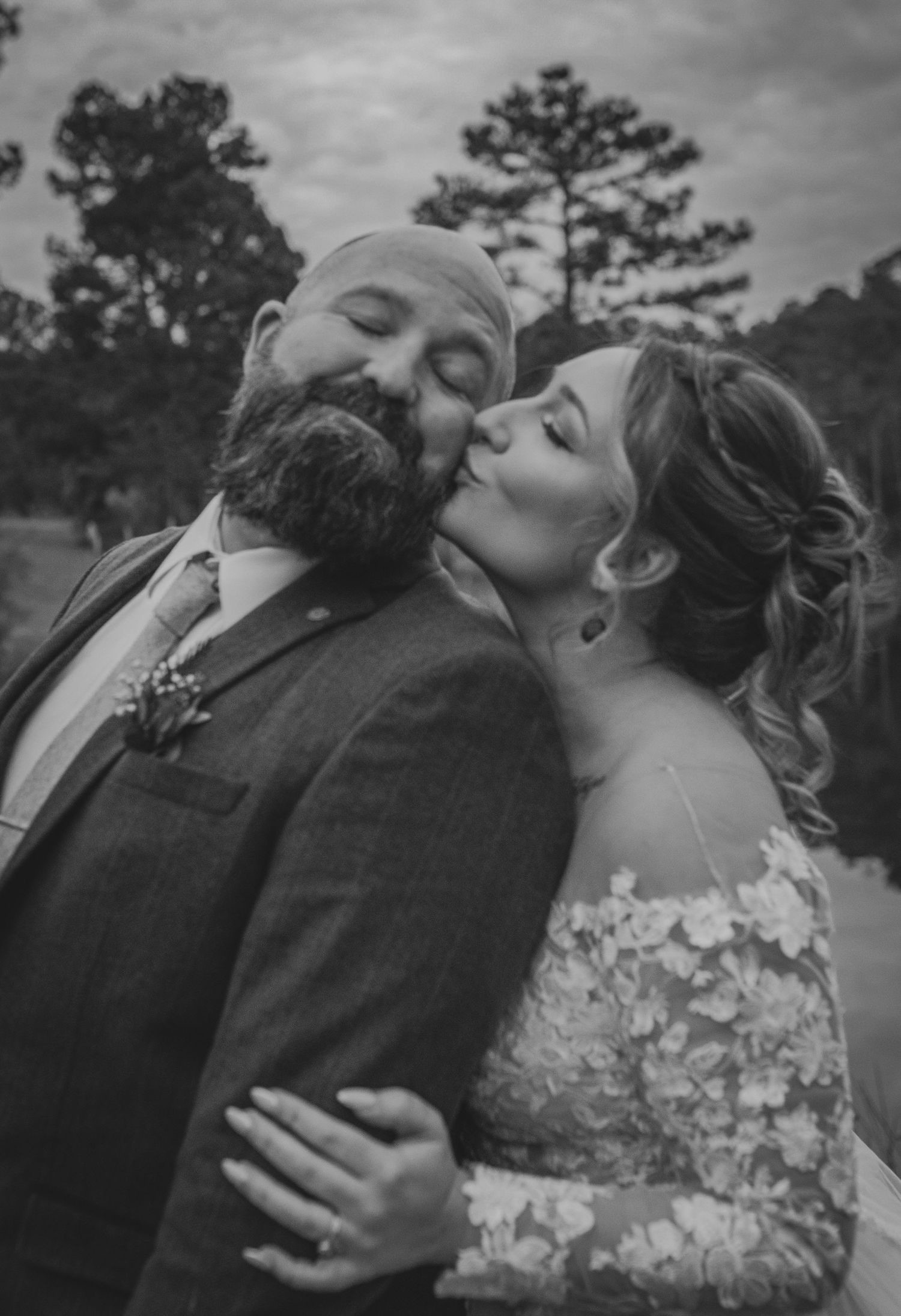 Black and white wedding photo of bride kissing groom on cheek, both dressed formally, outdoors among pine trees.