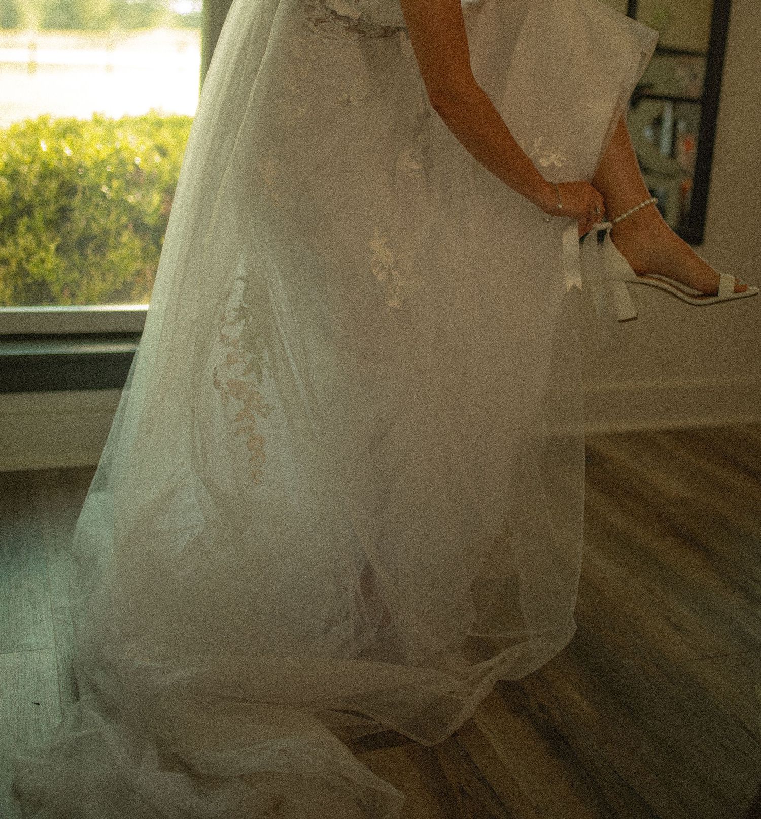 Bride in a white lace ball gown wedding dress with floral appliques, holding heels near a window.